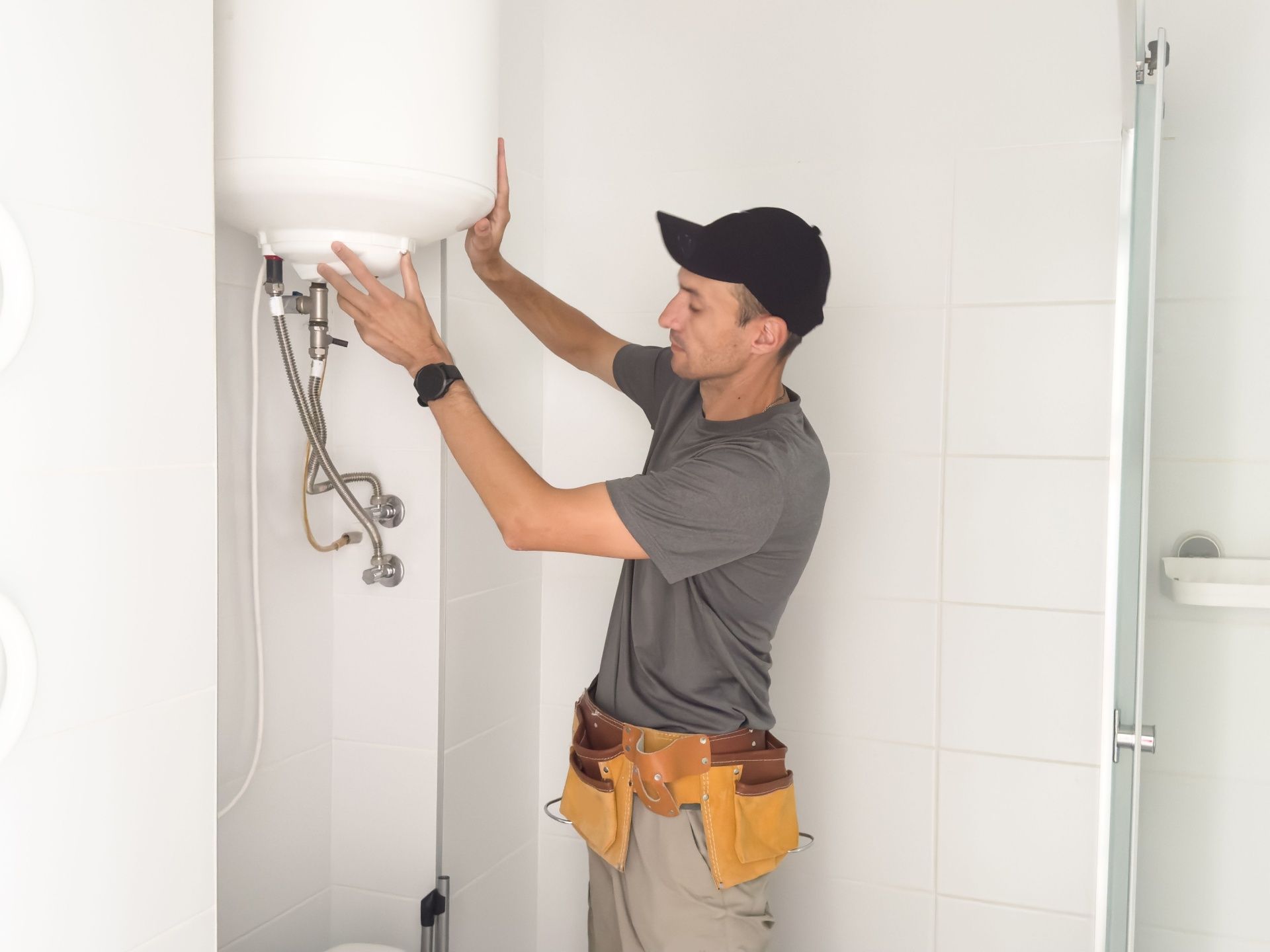 Man in cap with tool belt works on a water heater in a bathroom.