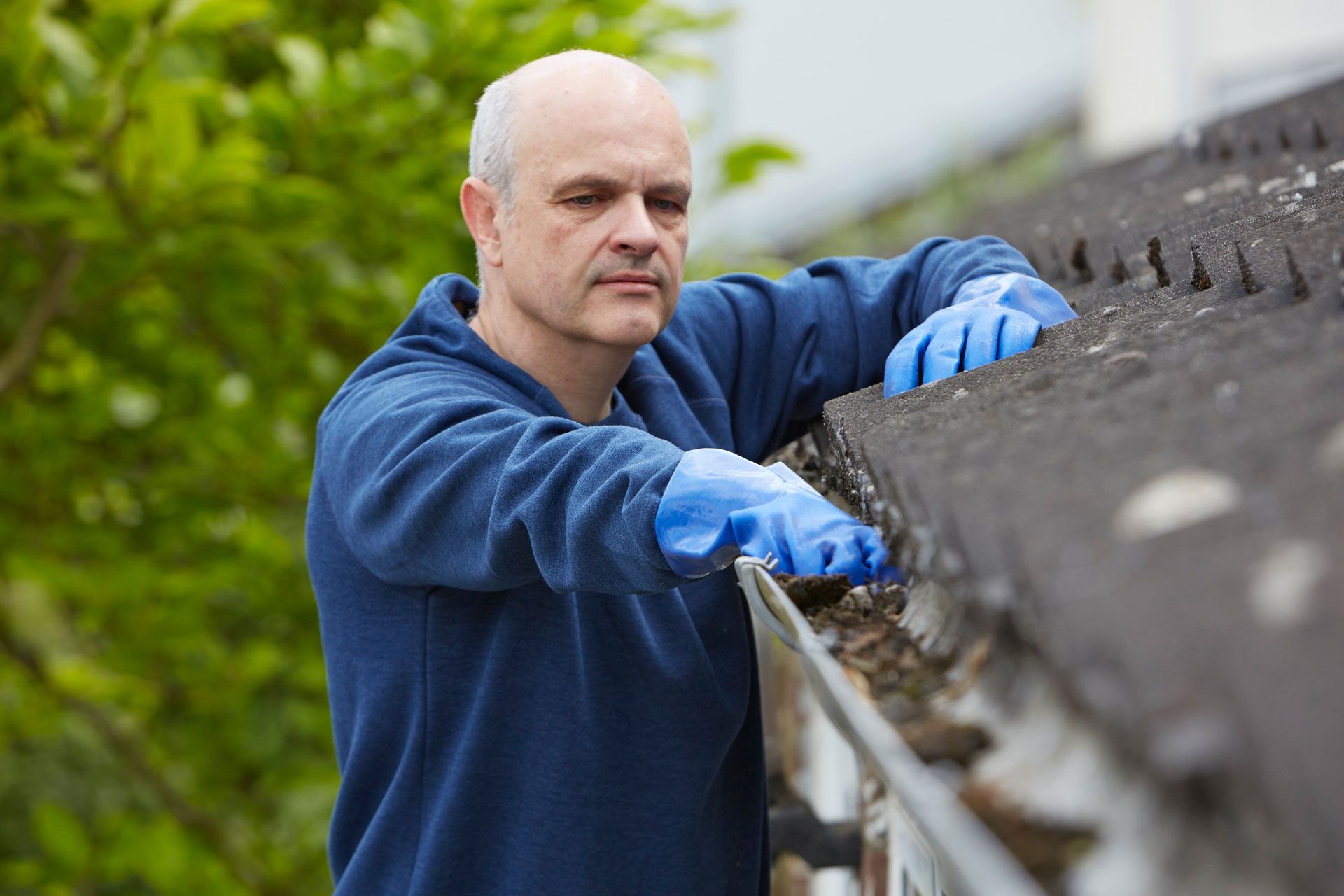 A man with gloves on a ladder doing gutter cleaning near trees under a blue sky.