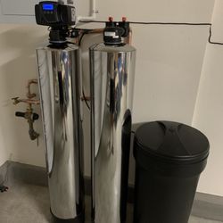 A water softener is sitting next to a black trash can in a room.