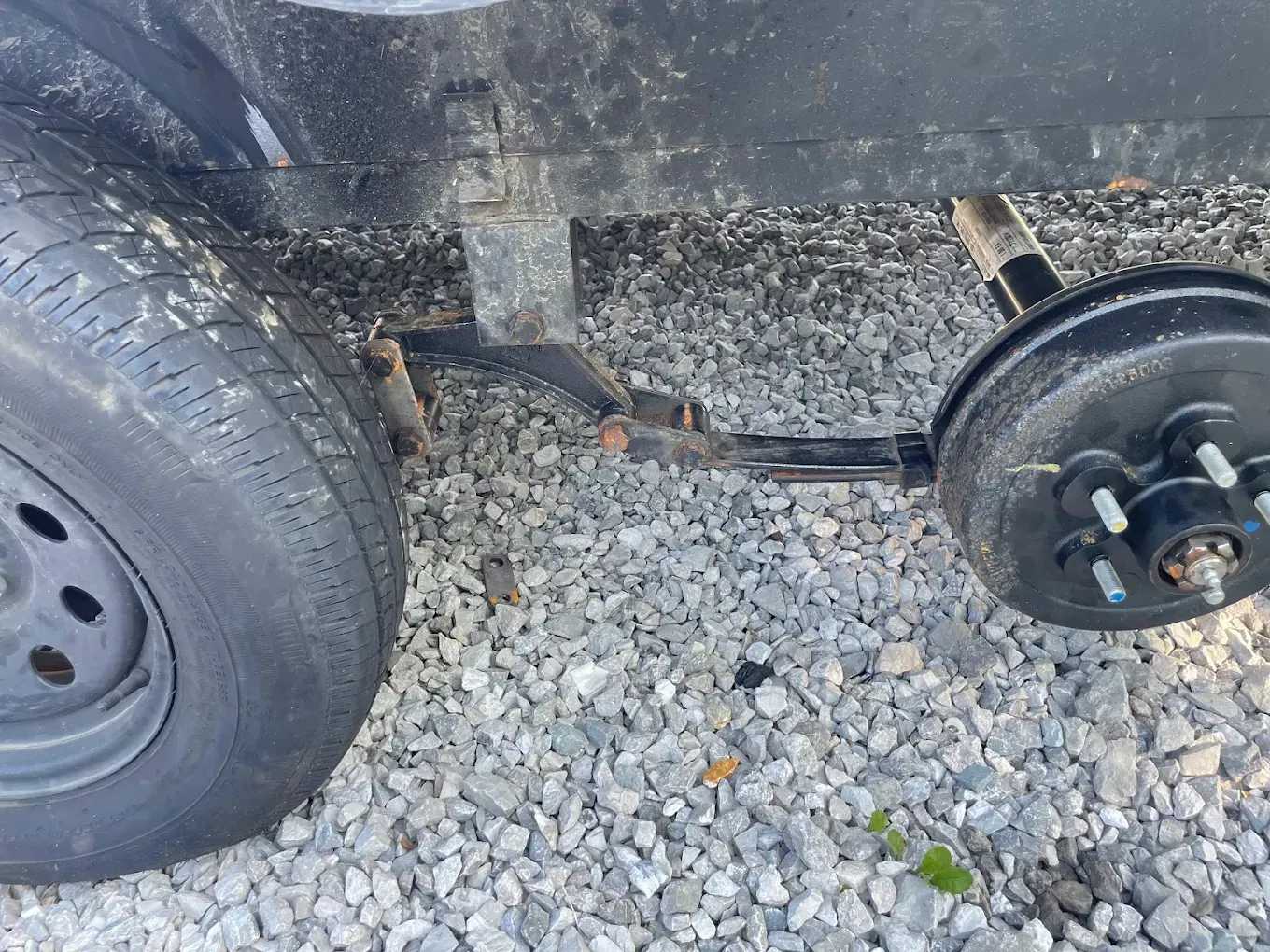 Black trailer with detached wheel. The axle is visible. Gravel ground.
