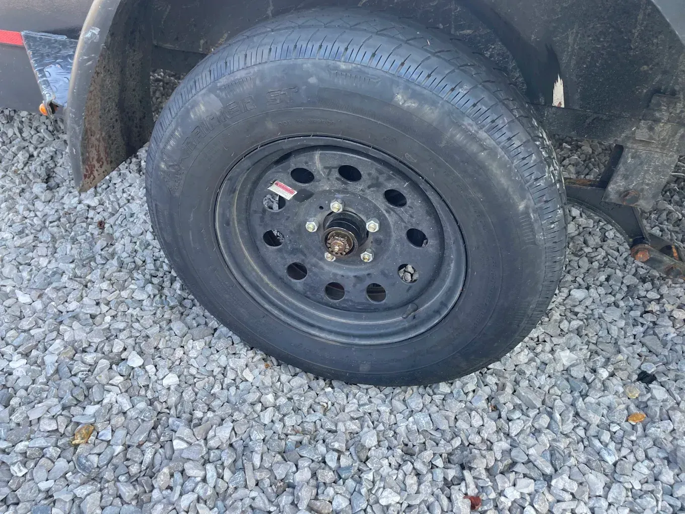 Black trailer tire on a steel rim, sitting on gravel.