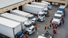 Trucks lined up at a loading dock with workers in orange vests; outdoor setting.