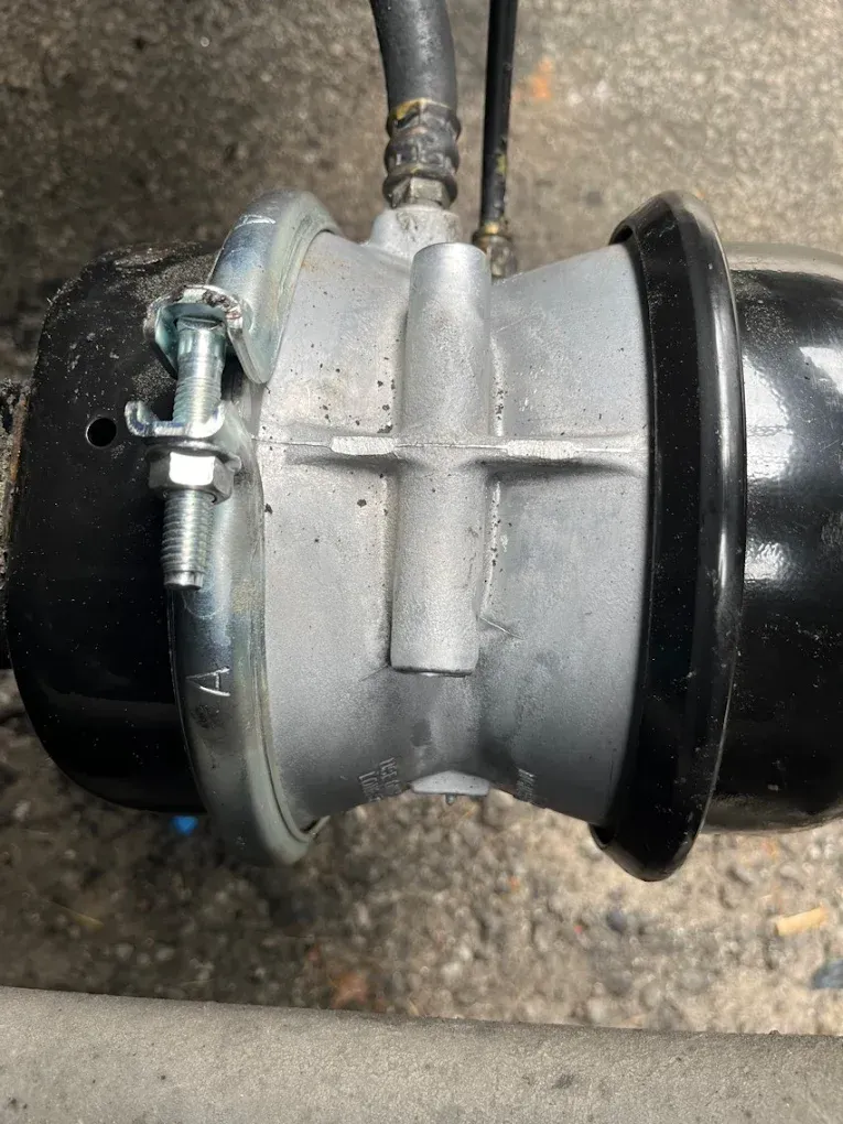 Air brake chamber on a vehicle, with a black rubber boot and metal housing, connected to hoses and a clamp.