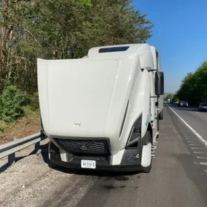 White semi-truck with hood open, parked on a road shoulder next to trees.