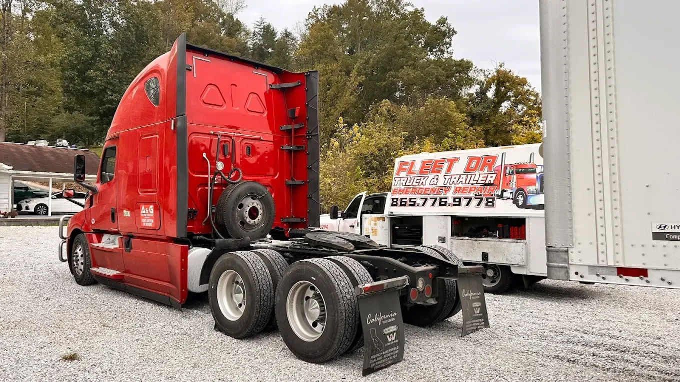 Red semi-truck with spare tire, parked next to a white trailer and service truck with a logo, outdoors.