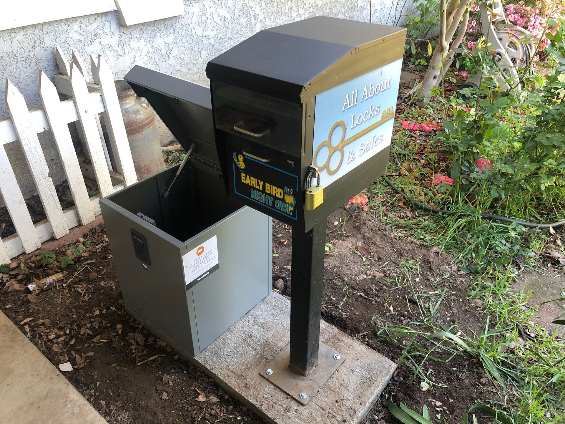 A mailbox with the lid open is sitting in the dirt next to a fence.