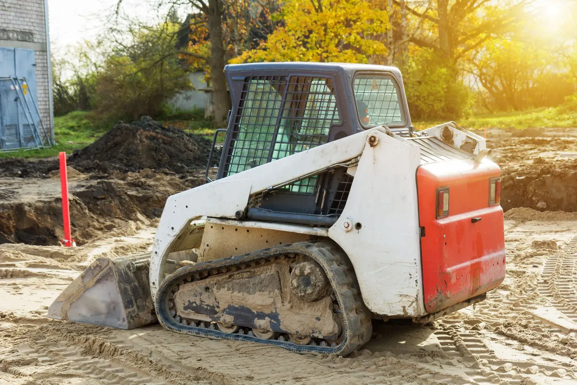 Small skid-steer loader with red rear panel working on a muddy construction site at sunset