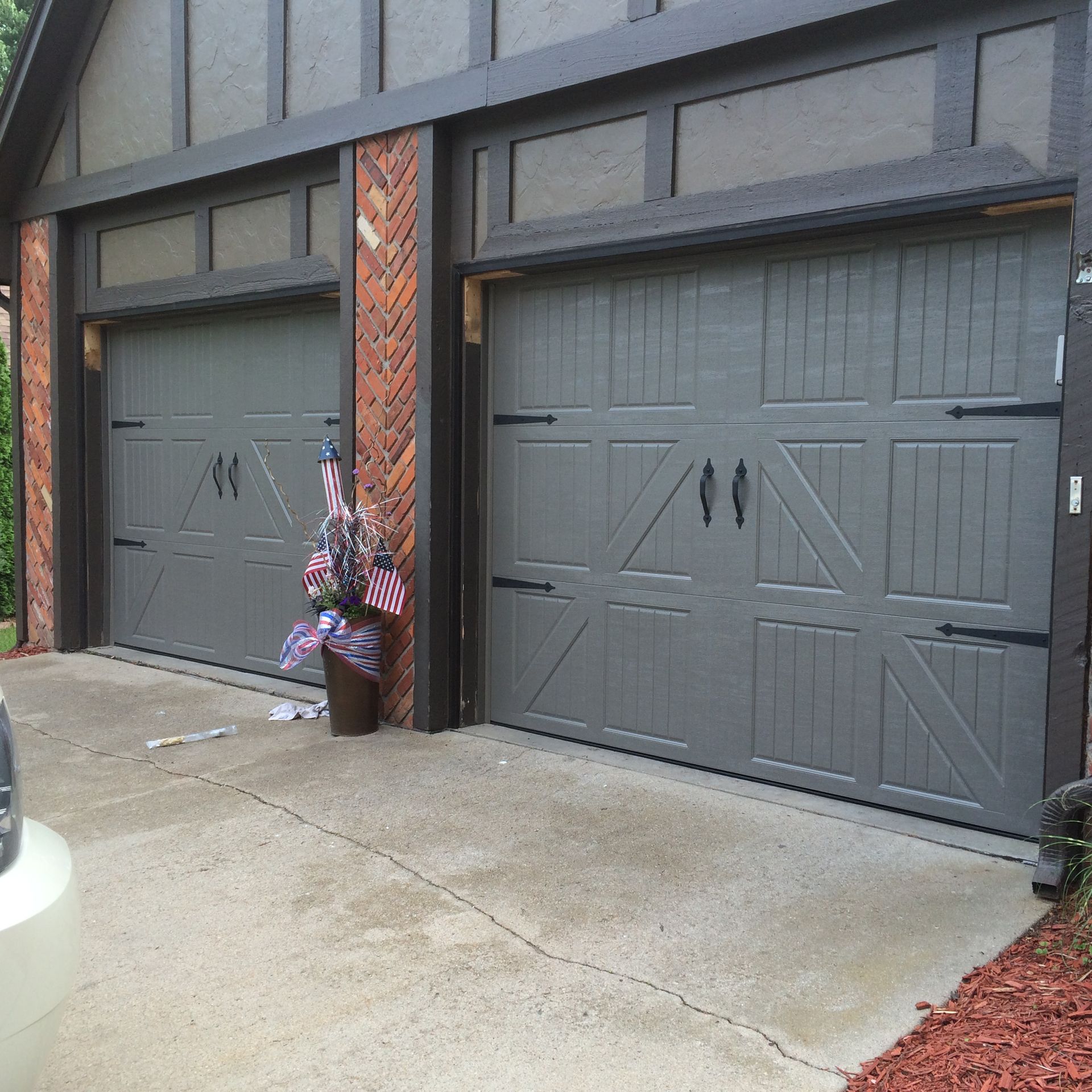 Two gray garage doors beside a wet driveway, with an umbrella and floral arrangement near the left door