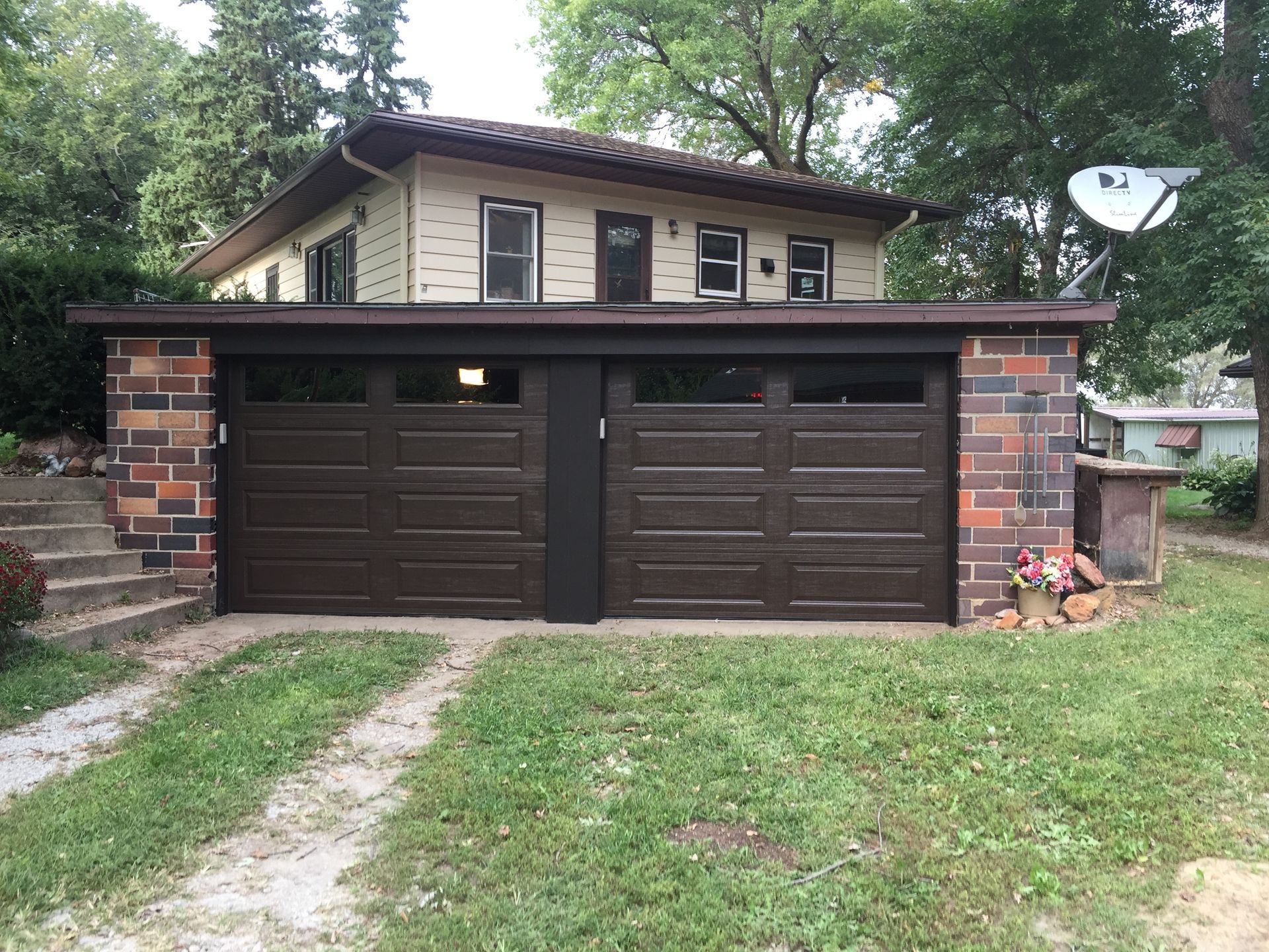 Two-car garage with dark brown doors and beige house above, viewed from a grassy driveway