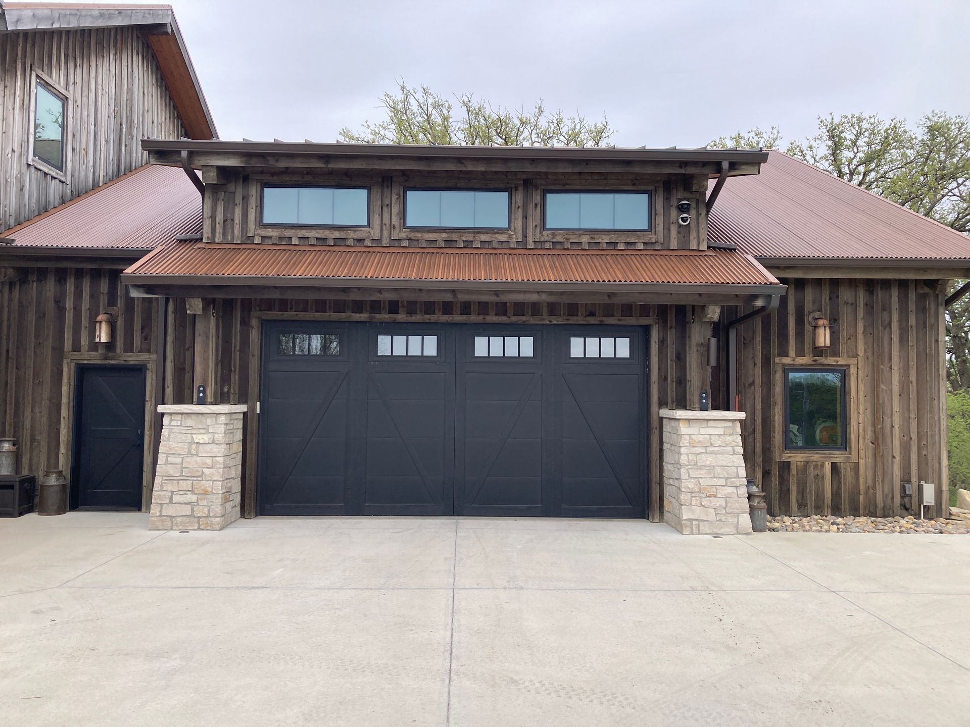 Rustic wooden building facade with a large black garage door and stone columns under a metal roof.