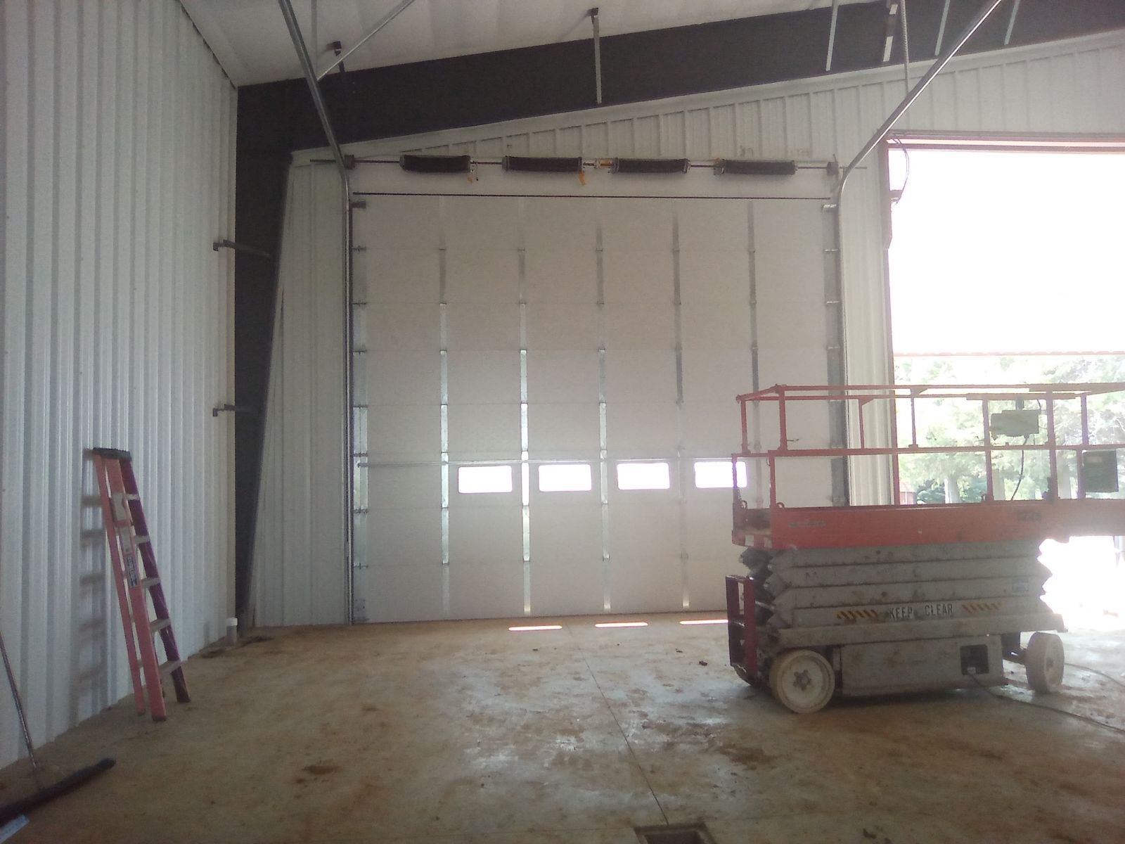 Empty warehouse interior with a large closed loading door, ladder on the left, and a scissor lift on the right.