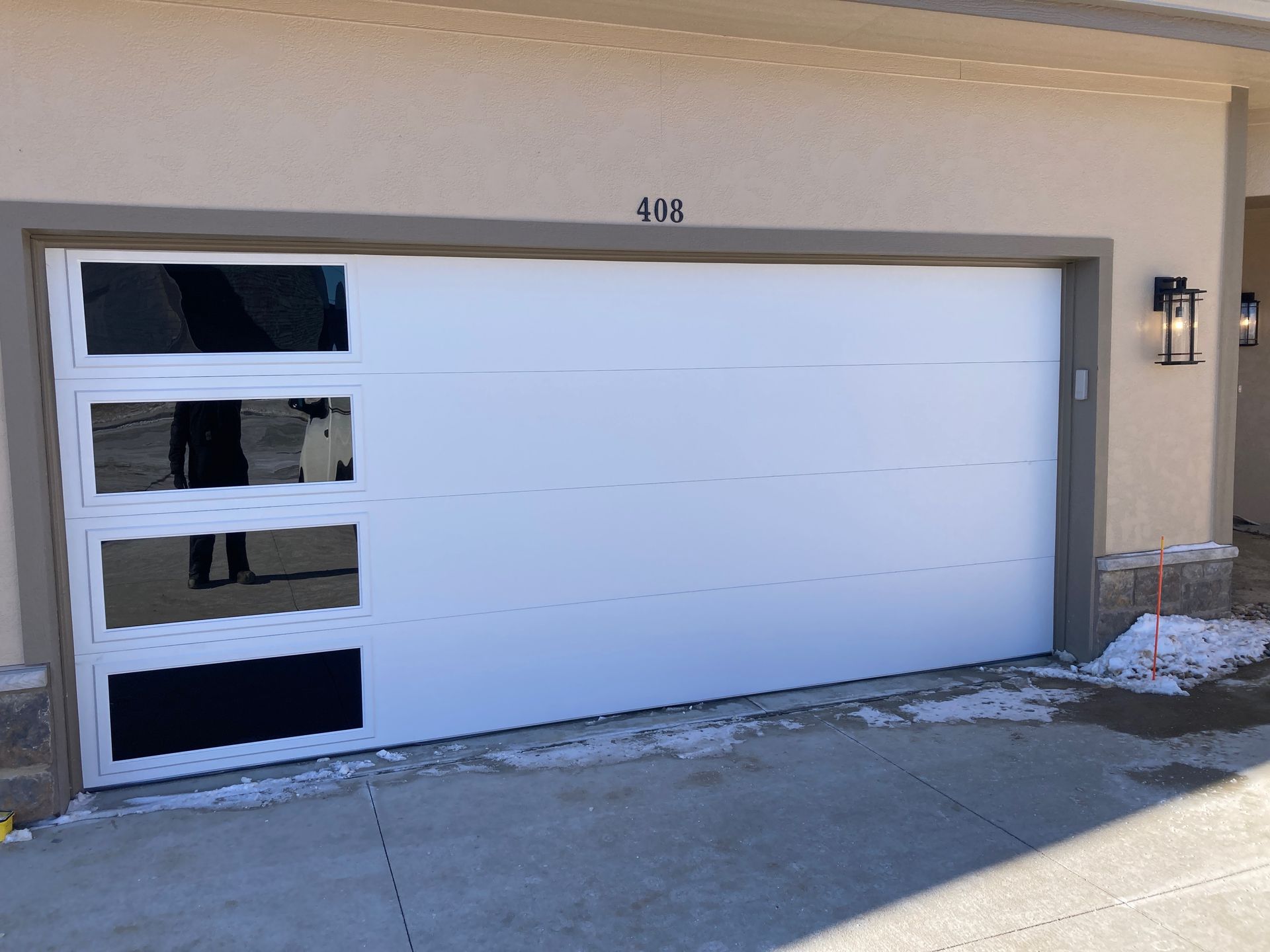White garage door with three black window panels beside a house entrance and wall-mounted light.