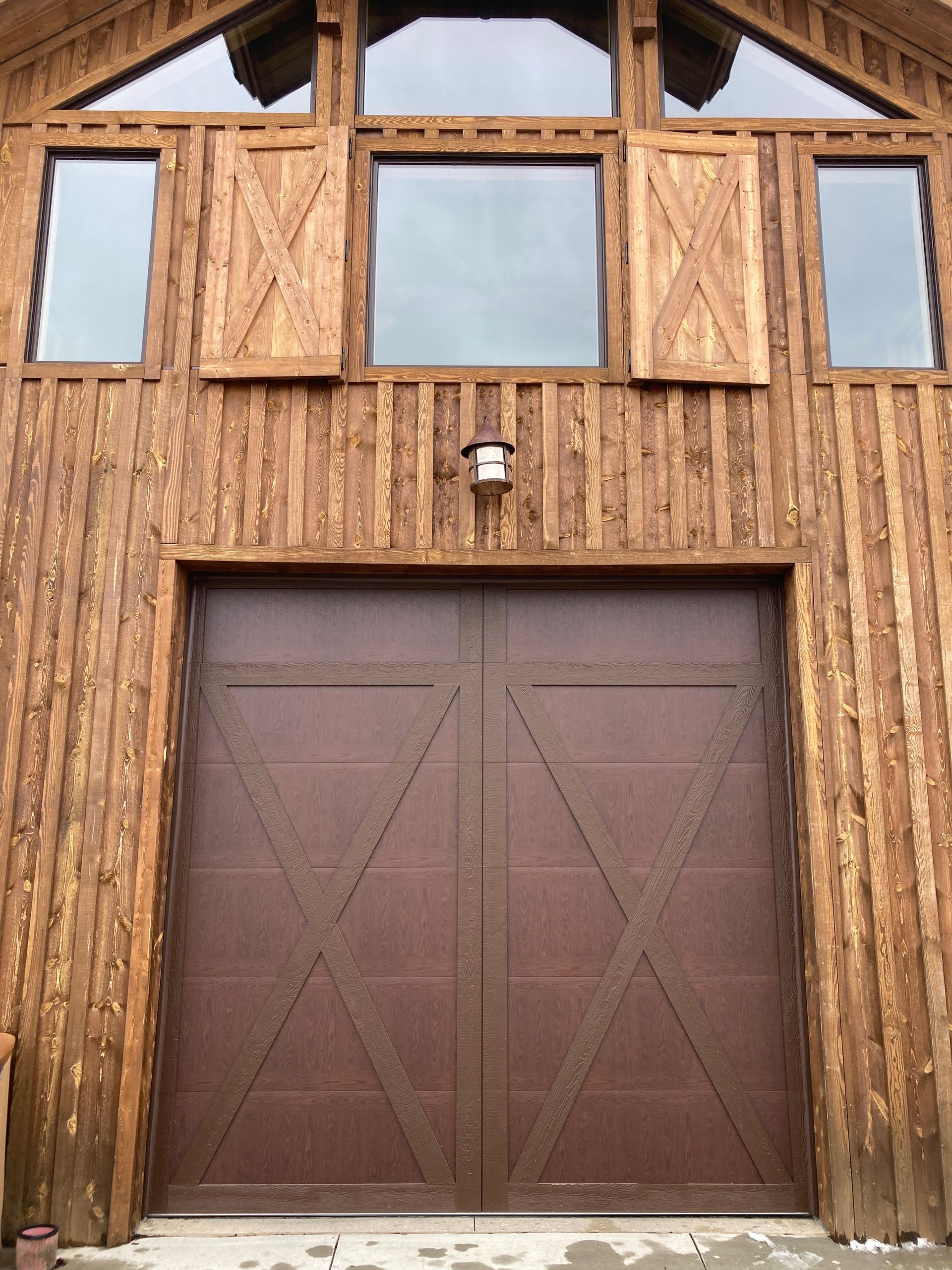 Rustic wooden garage door beneath a timber facade with small windows and a central light fixture