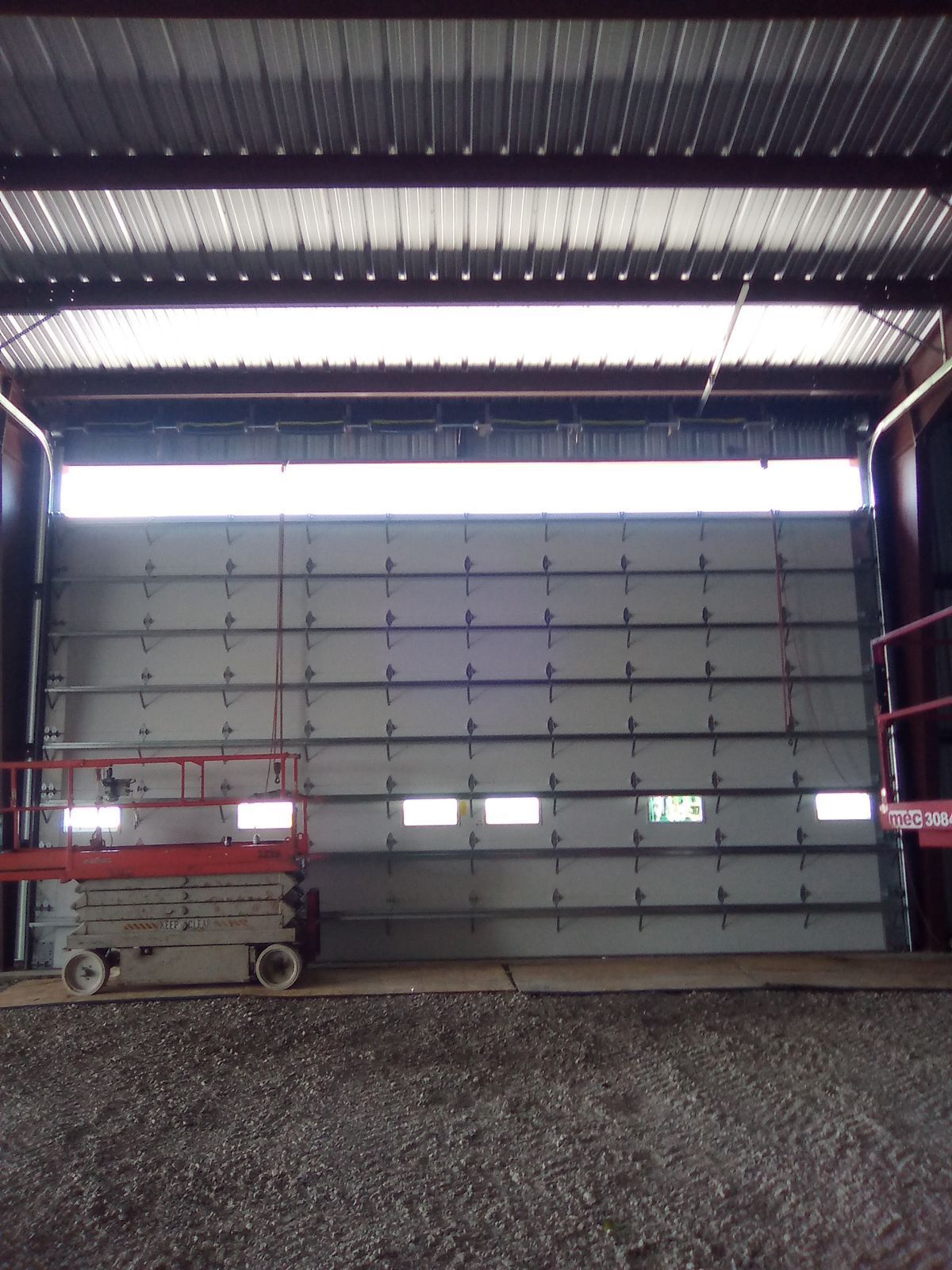 Large closed industrial garage door inside a warehouse, with a scissor lift on the left and gravel floor.