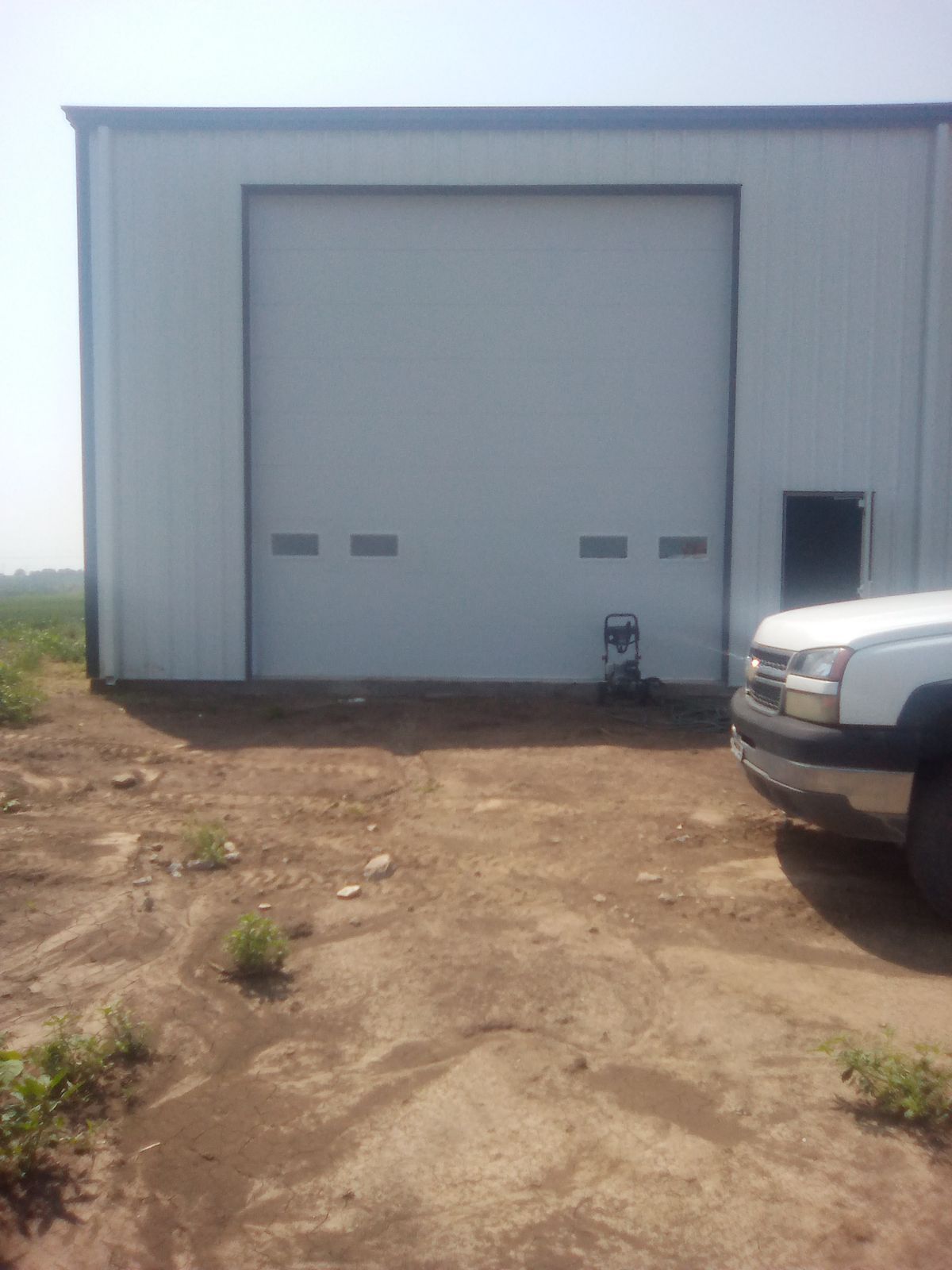 Gray metal warehouse with a large roll-up door, dirt lot in front, and part of a white truck at right.