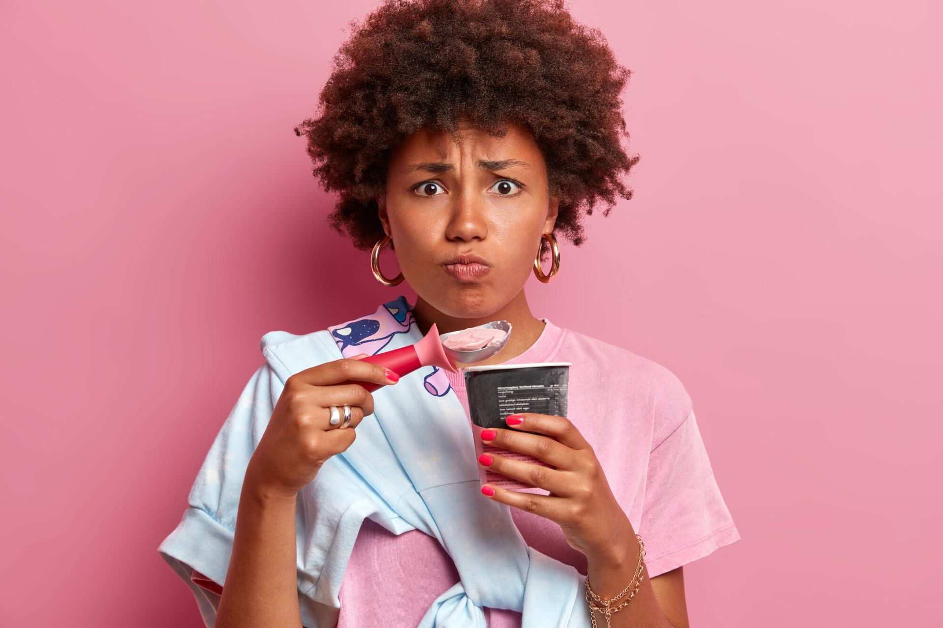 A person with curly hair, wearing a pink shirt, holds an ice cream scoop and cup while making a pouty facial expression.