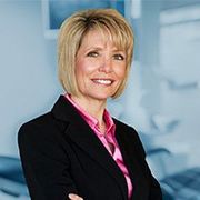 A woman in a black suit and pink shirt is smiling with her arms crossed.