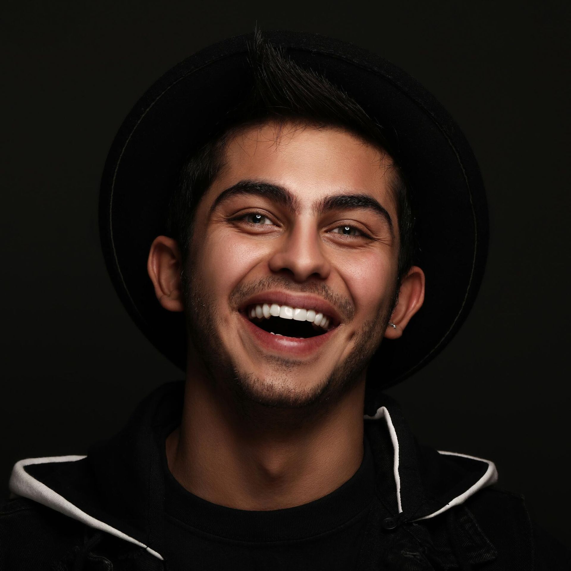 A young man wearing a hat is laughing with his mouth open.