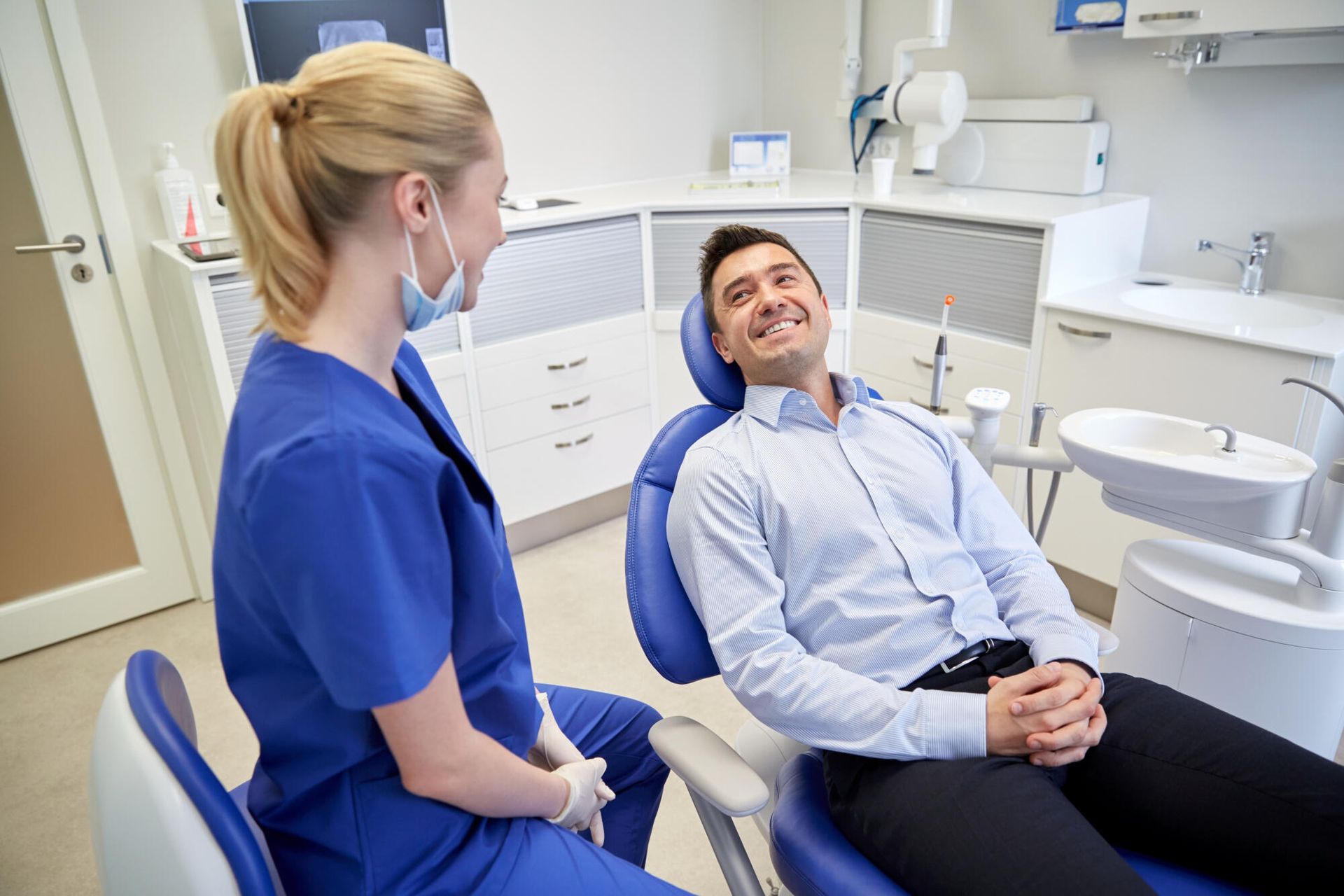 Dentist in blue scrubs talking to a smiling male patient in a dental chair.