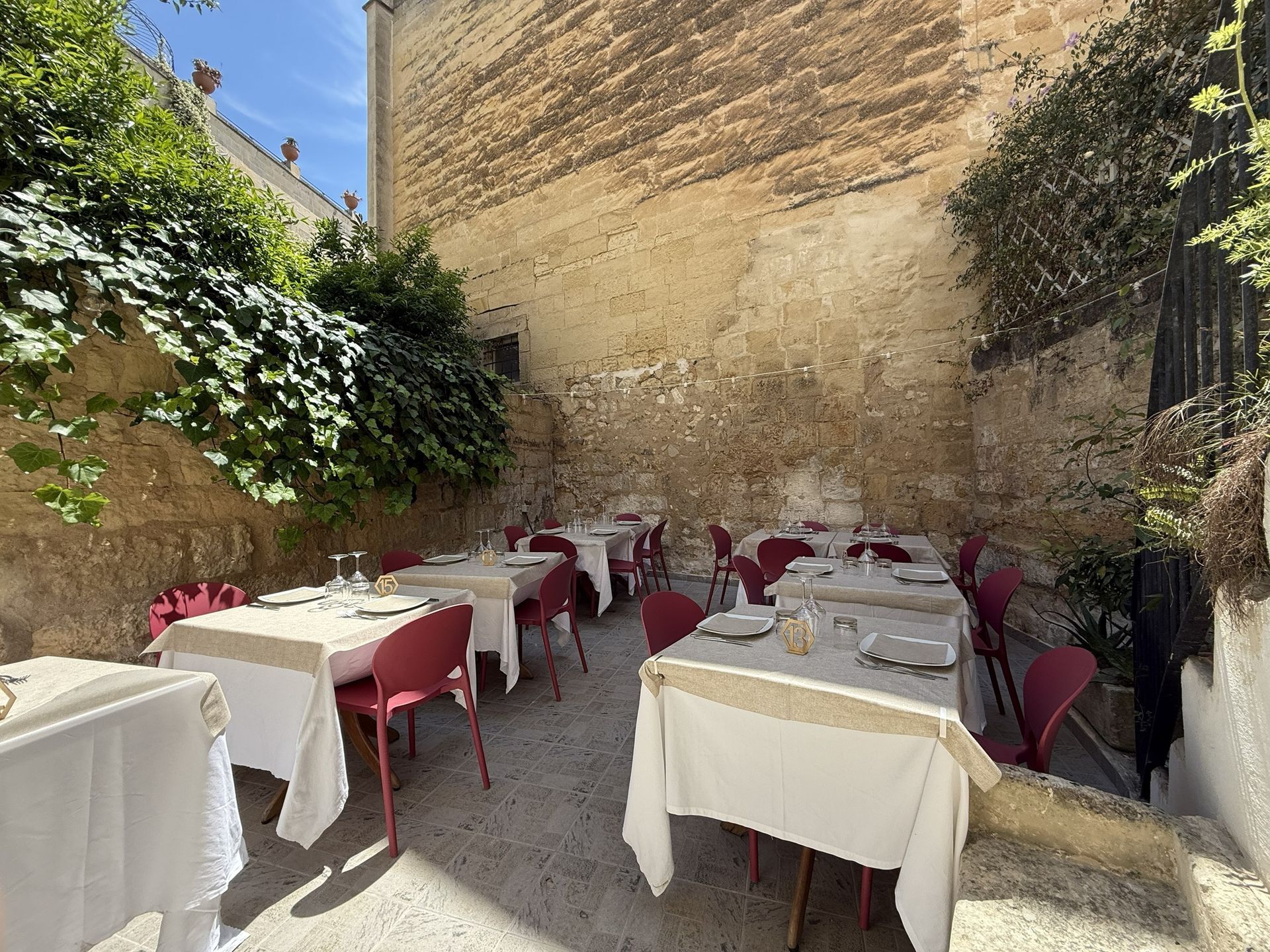 Terrazza esterna del ristorante con sedie rosse e tovaglie bianche accanto a un muro di pietra e vegetazione.