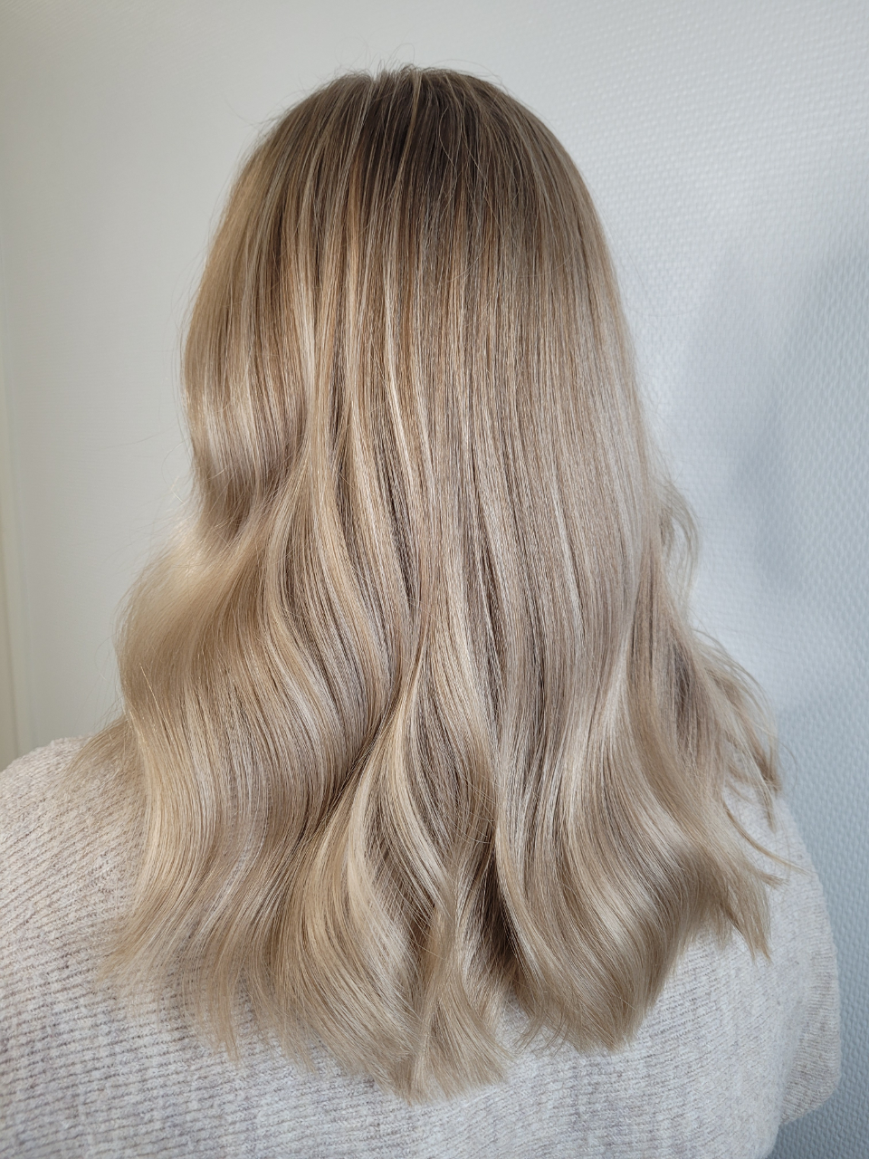 Blonde hair styled with crimped texture and gentle waves, over a light sweater against a white wall.
