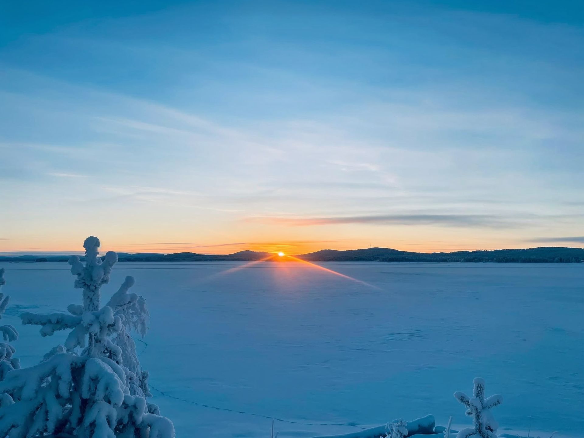 Sunrise over a snow-covered landscape with a frozen lake and snow-laden trees under a blue sky.