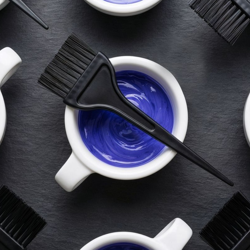 Hair dye bowl with blue dye and applicator brush, surrounded by other bowls and brushes on black surface.