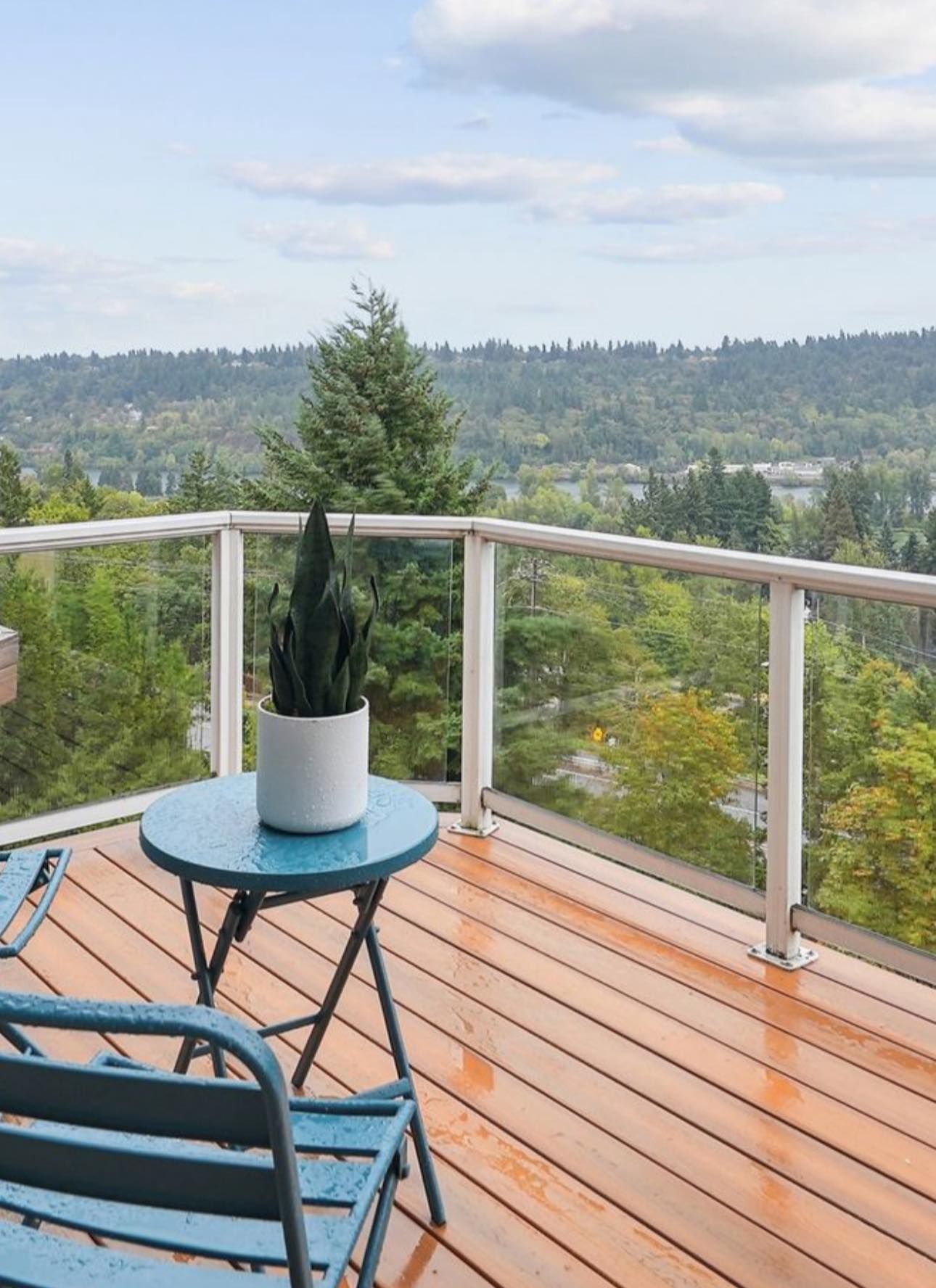 A blue bistro table and chair on a wet wooden deck overlooking a lush, forested valley under a cloudy sky.