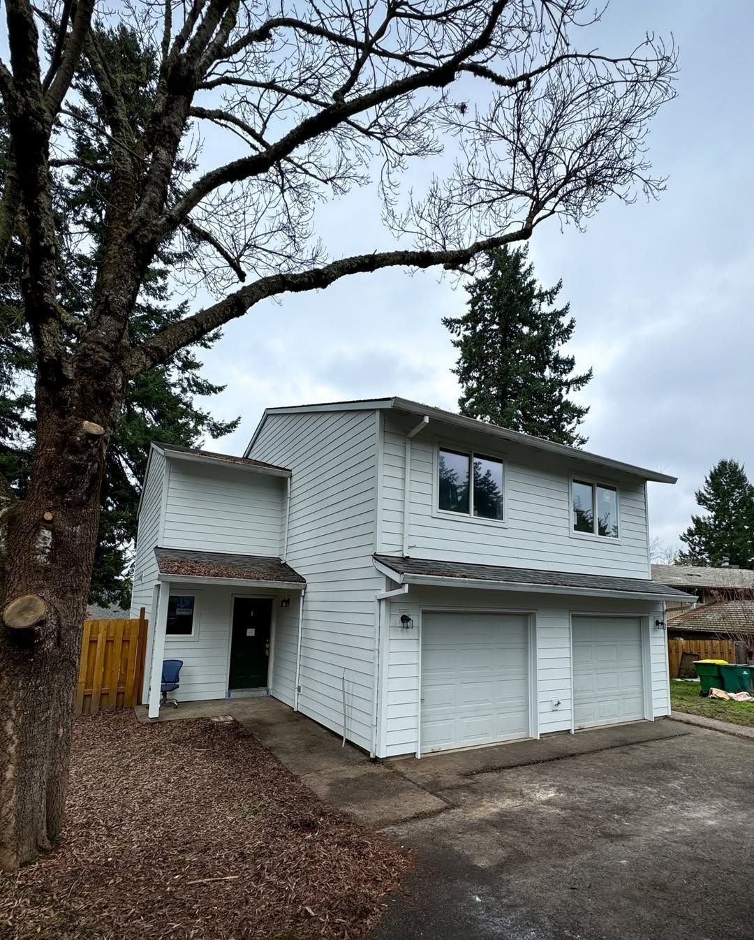 A white two-story house with a two-car garage and a small front entrance under a large, leafless tree.