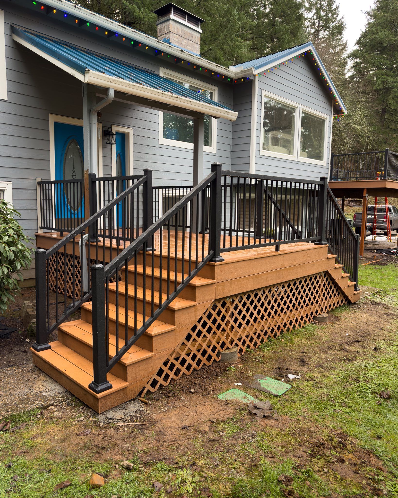 A newly constructed wooden deck with black railings and lattice skirting stands outside a gray house with a blue door.