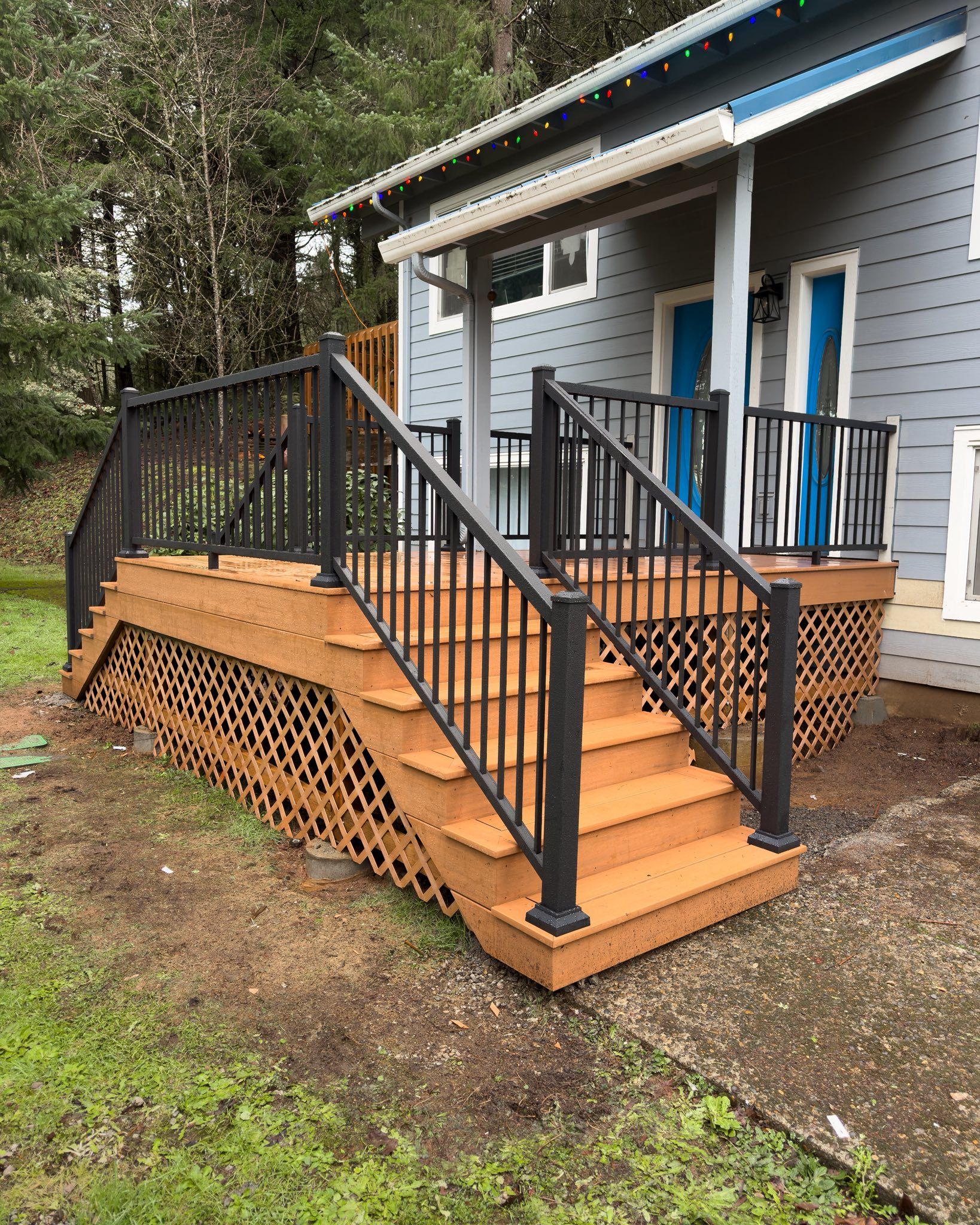 A wooden deck with a staircase and black metal railings sits beside a house, finished with a lattice skirt.