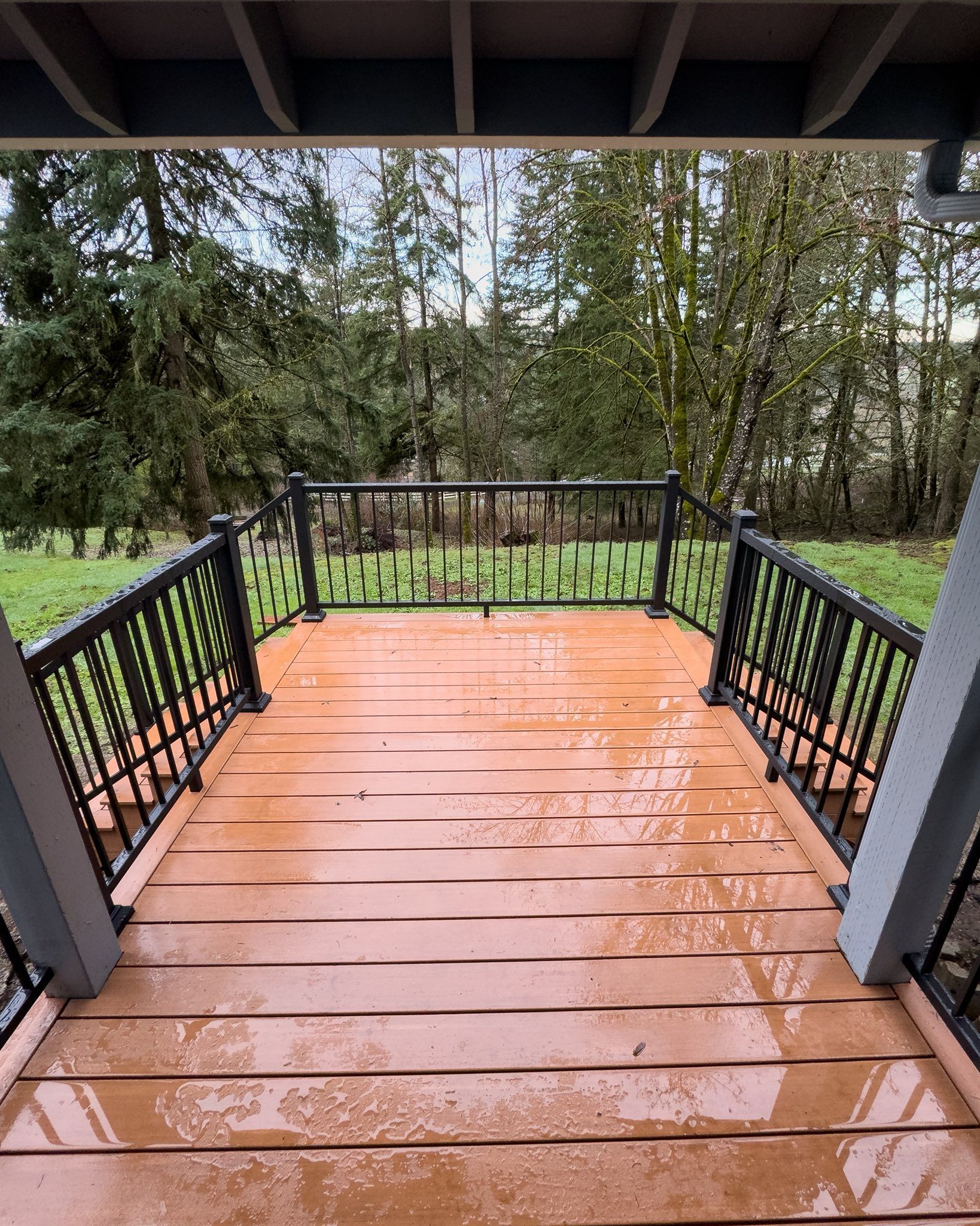 A wooden deck with black railings looks out onto a green, tree-filled yard on a wet, rainy day.