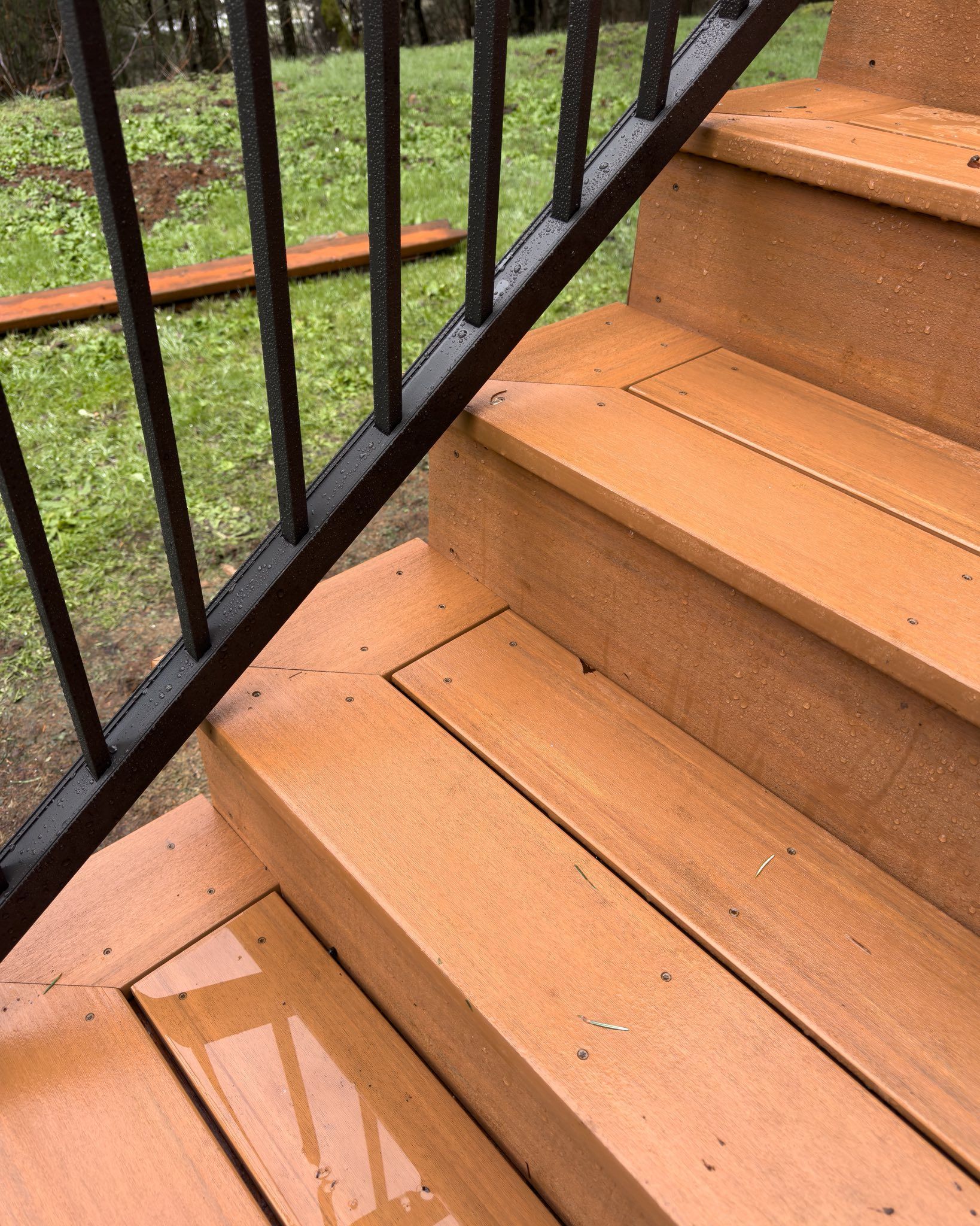 A close-up view of wooden outdoor stairs with a black metal handrail, wet from rain, leading down toward a grassy yard.