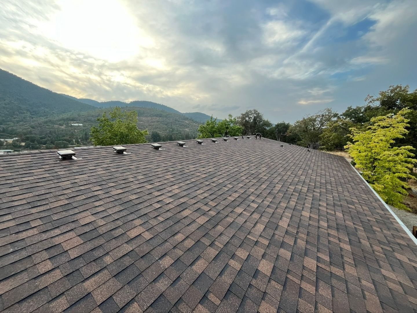 A brown, asphalt-shingled roof under a cloudy sky, with several roof vents visible along the ridgeline and a scenic backdrop.