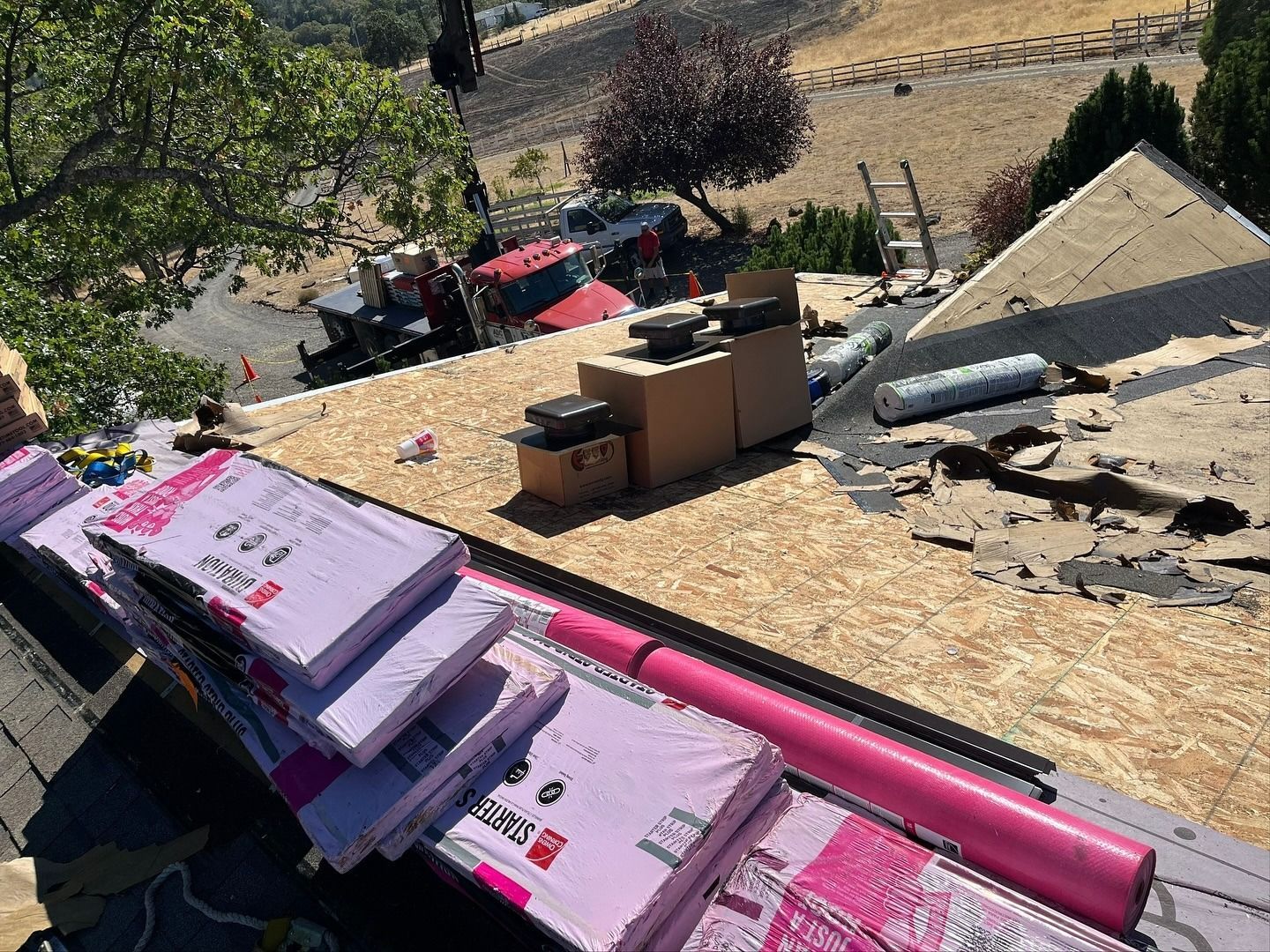 Construction materials including purple foam board insulation and a pink roll rest on a plywood-sheathed residential roof.