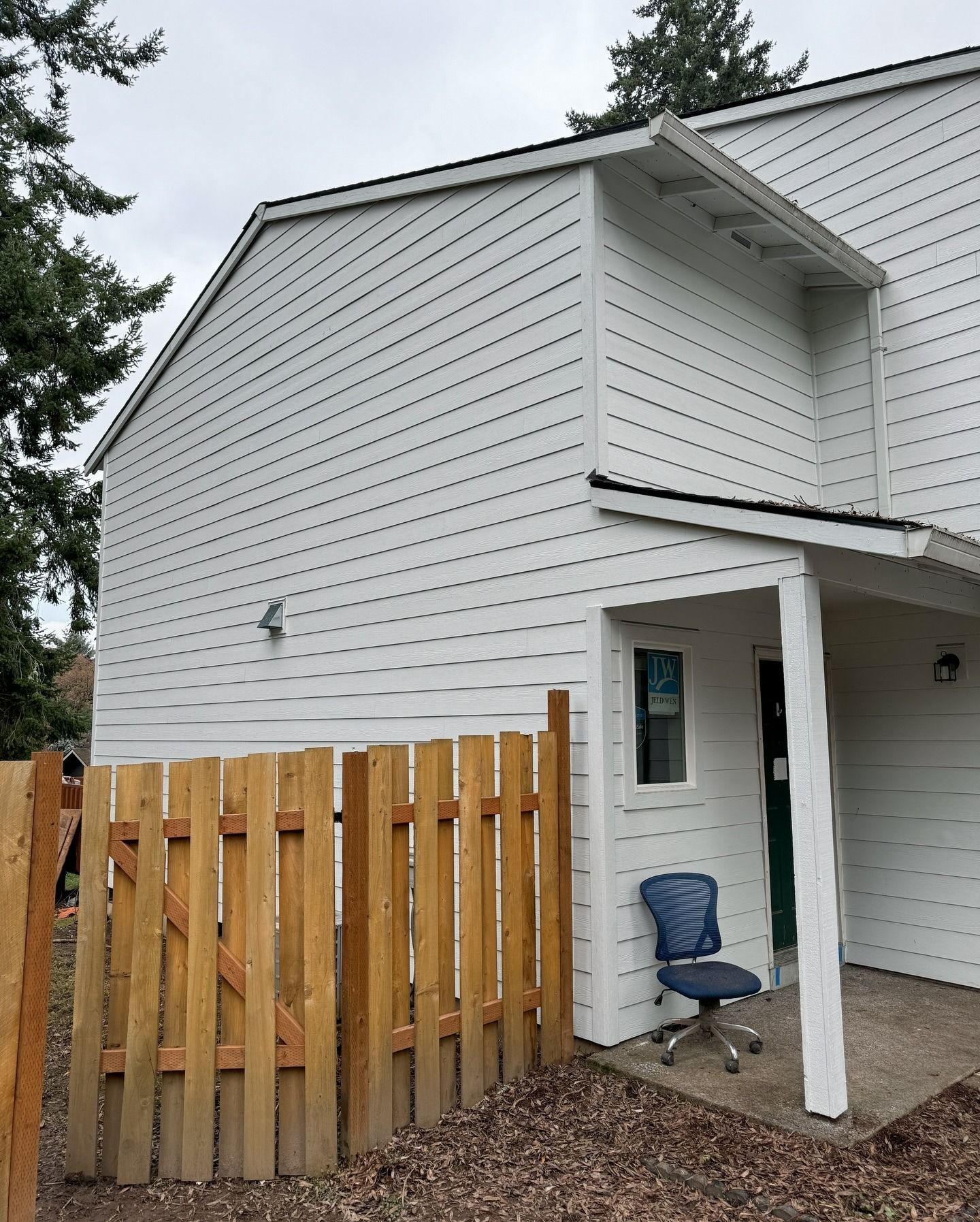 A blue office chair sits on a concrete patio next to a wooden fence and the white-sided wall of a building.