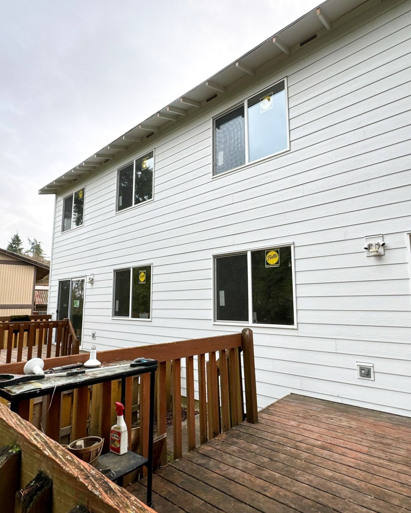 A white, two-story house exterior with siding and new windows viewed from a wooden deck under a cloudy sky.