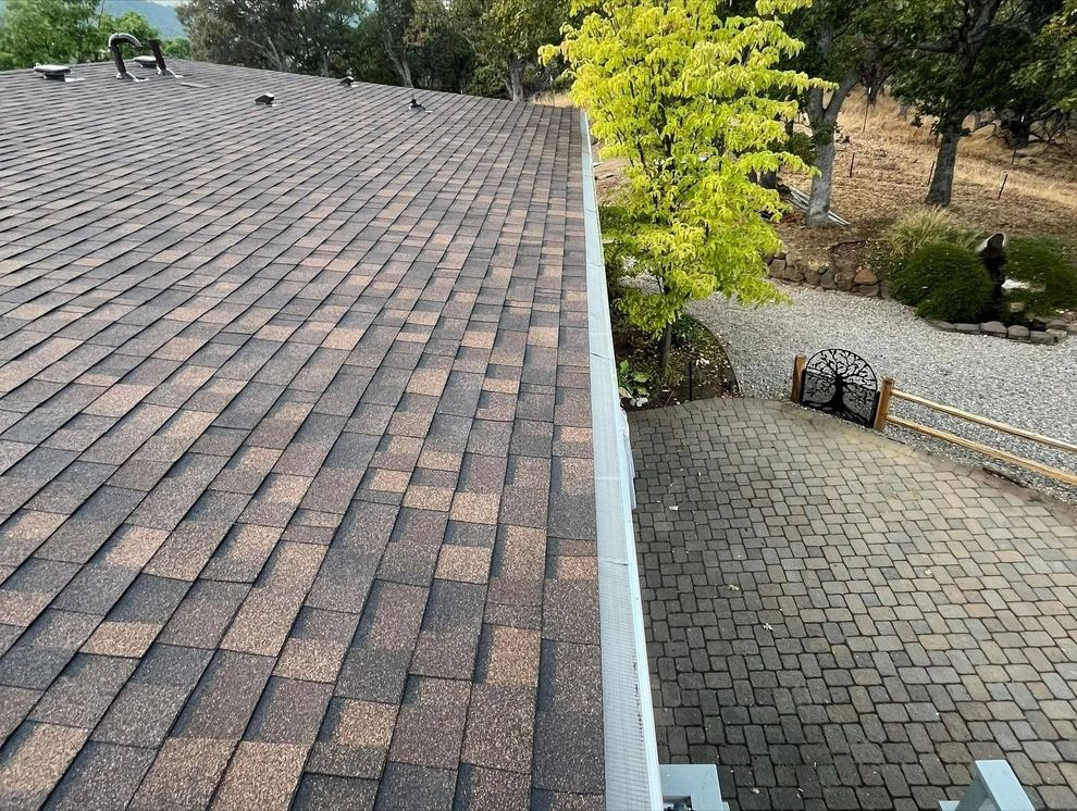 A high-angle view of a brown asphalt shingle roof with a white gutter guard, overlooking a stone driveway and trees.