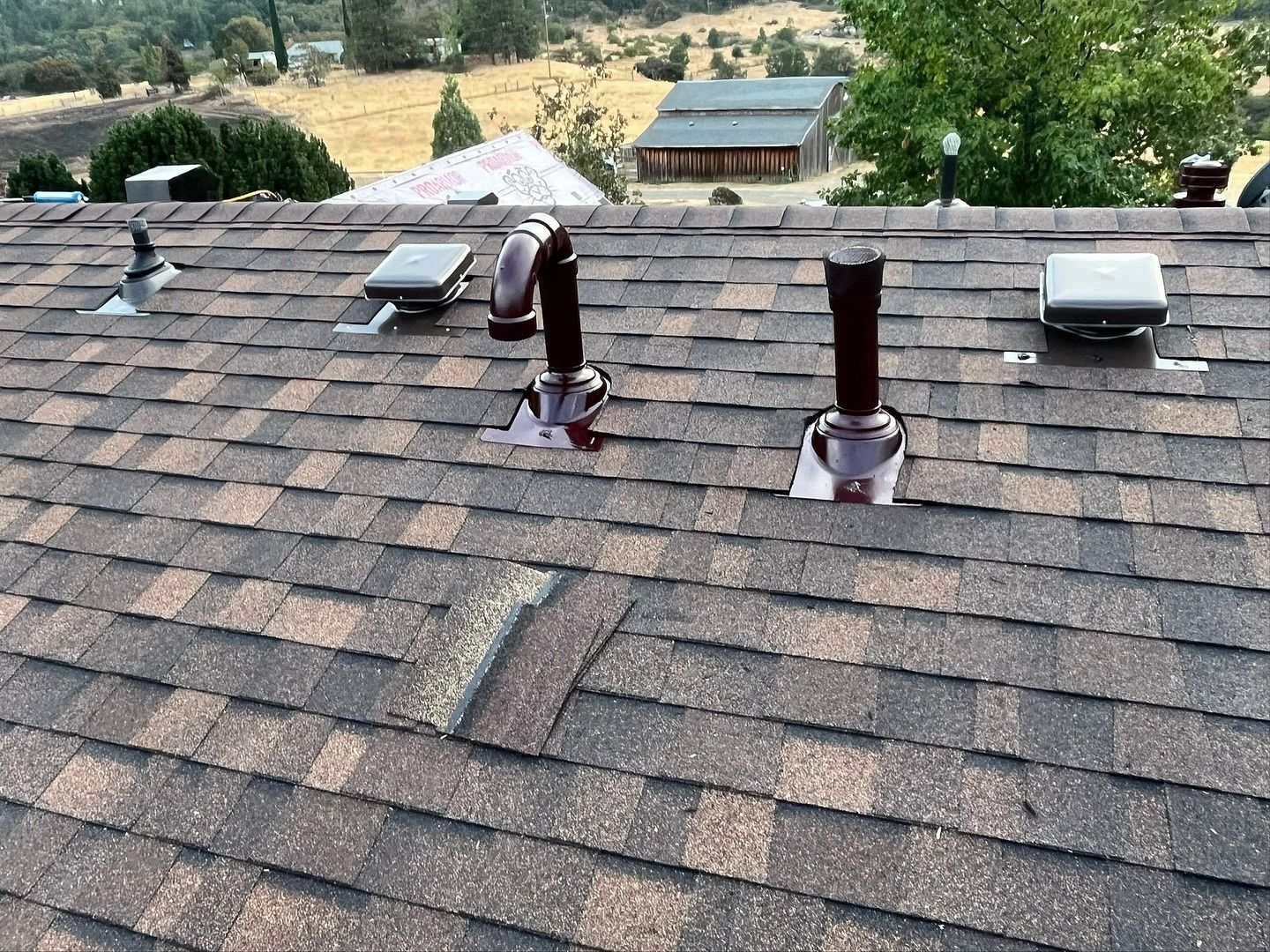 A residential shingled roof with several metal plumbing vents and square roof vents, overlooking a rural landscape.