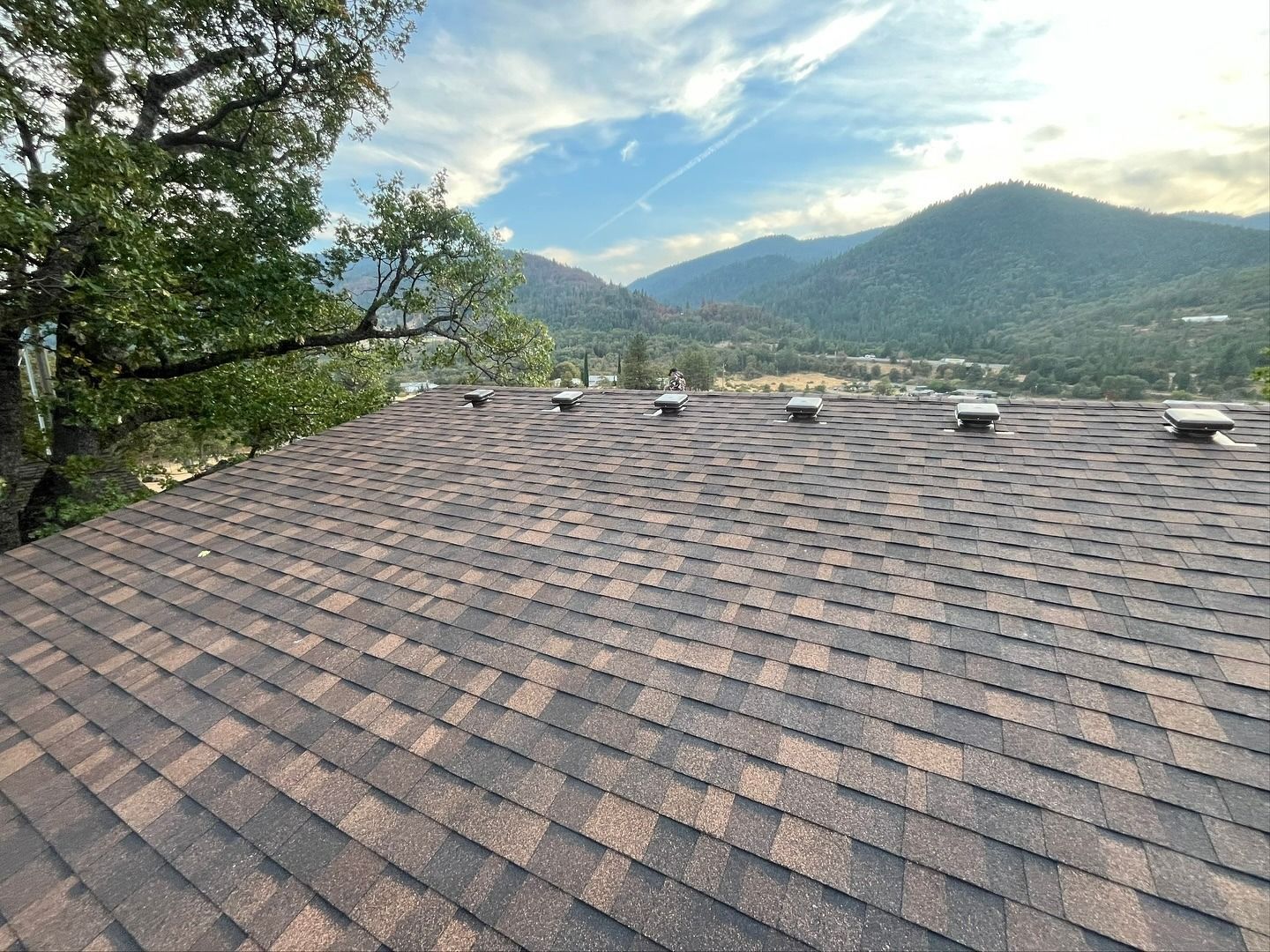 A brown asphalt shingle roof with several vents, overlooking a scenic mountain valley under a partly cloudy sky.