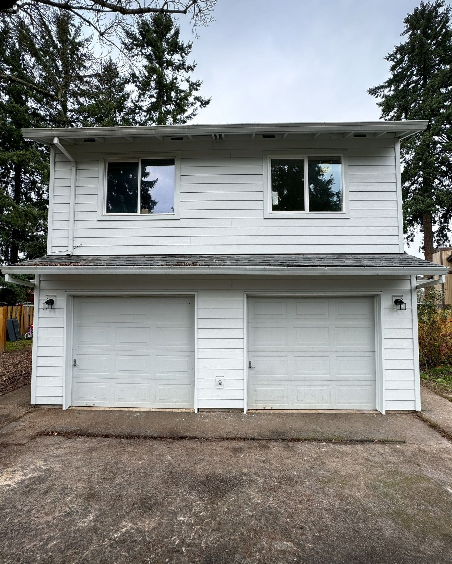 A white two-story detached garage with two garage doors and two upper-level windows, situated outdoors.