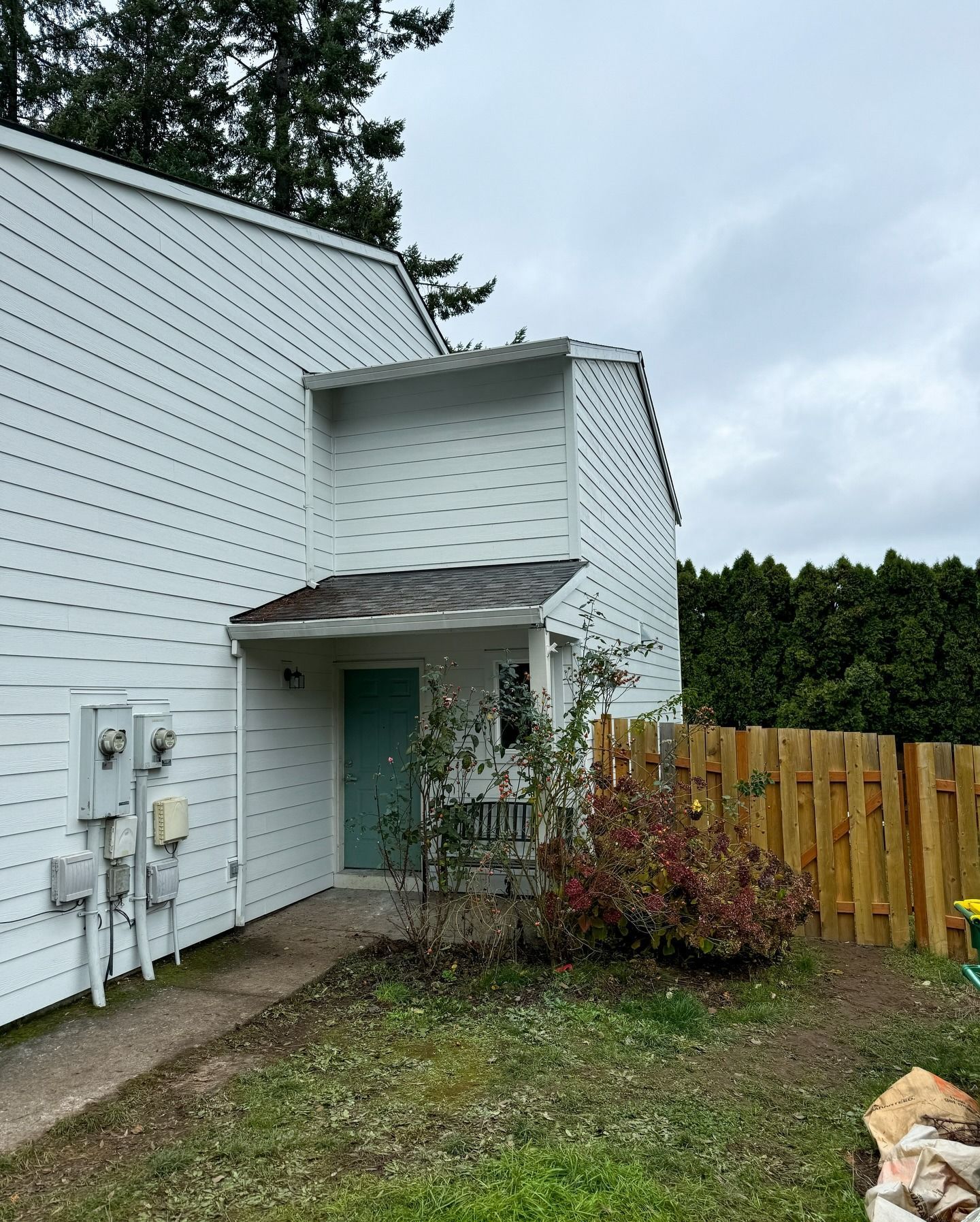 A white, two-story residential building with a teal front door, small porch, and wooden fence on an overcast day.