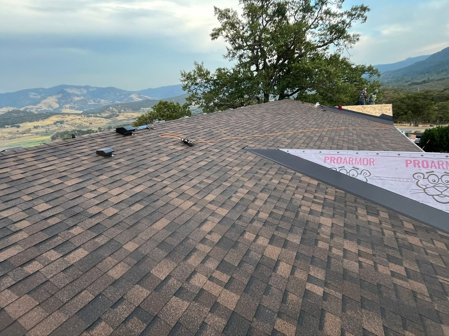 A roof covered in brown asphalt shingles with a section of pink underlayment, overlooking a scenic mountain landscape.