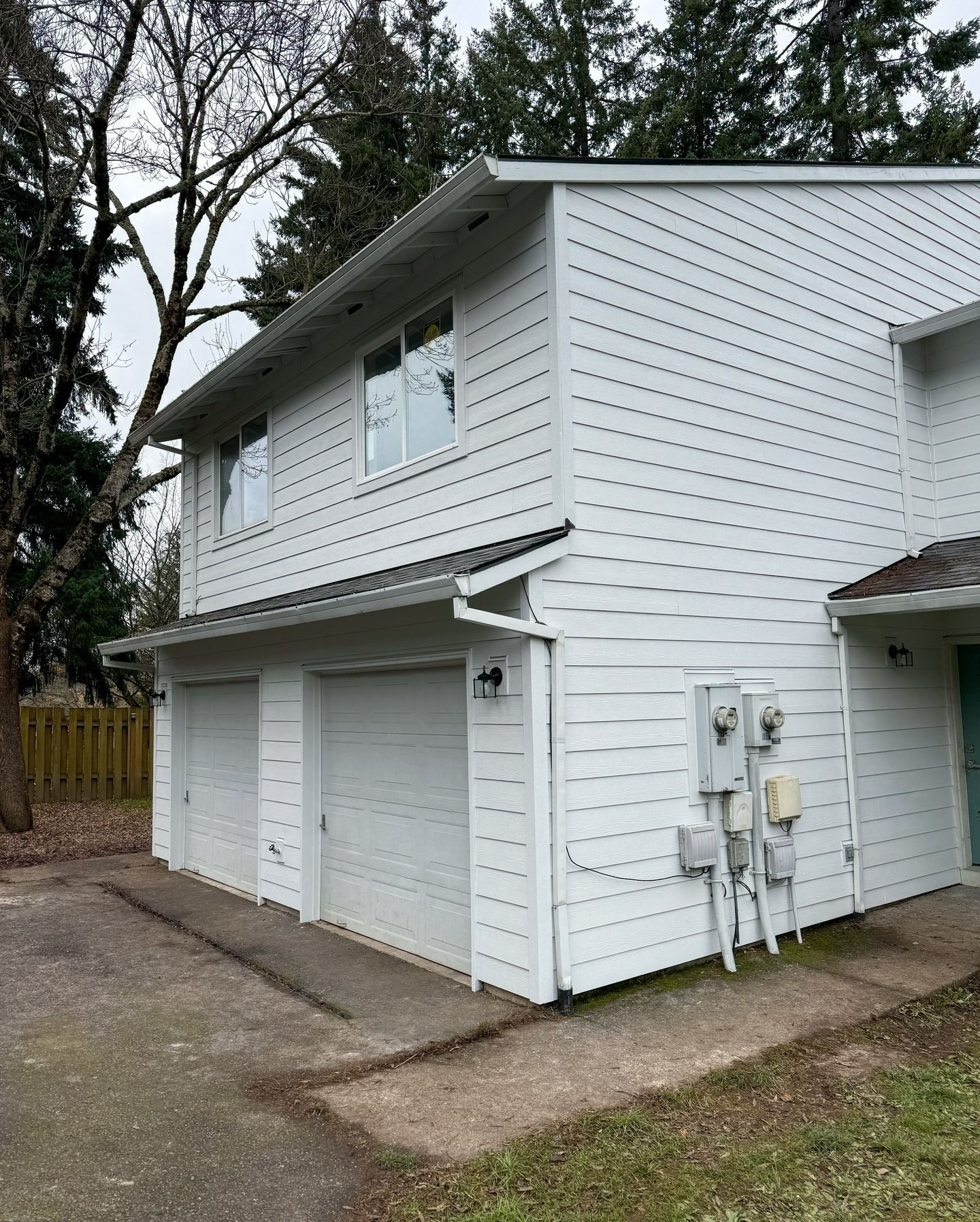 A two-story white building with a dual-car garage and a small paved driveway, set against a backdrop of bare trees.