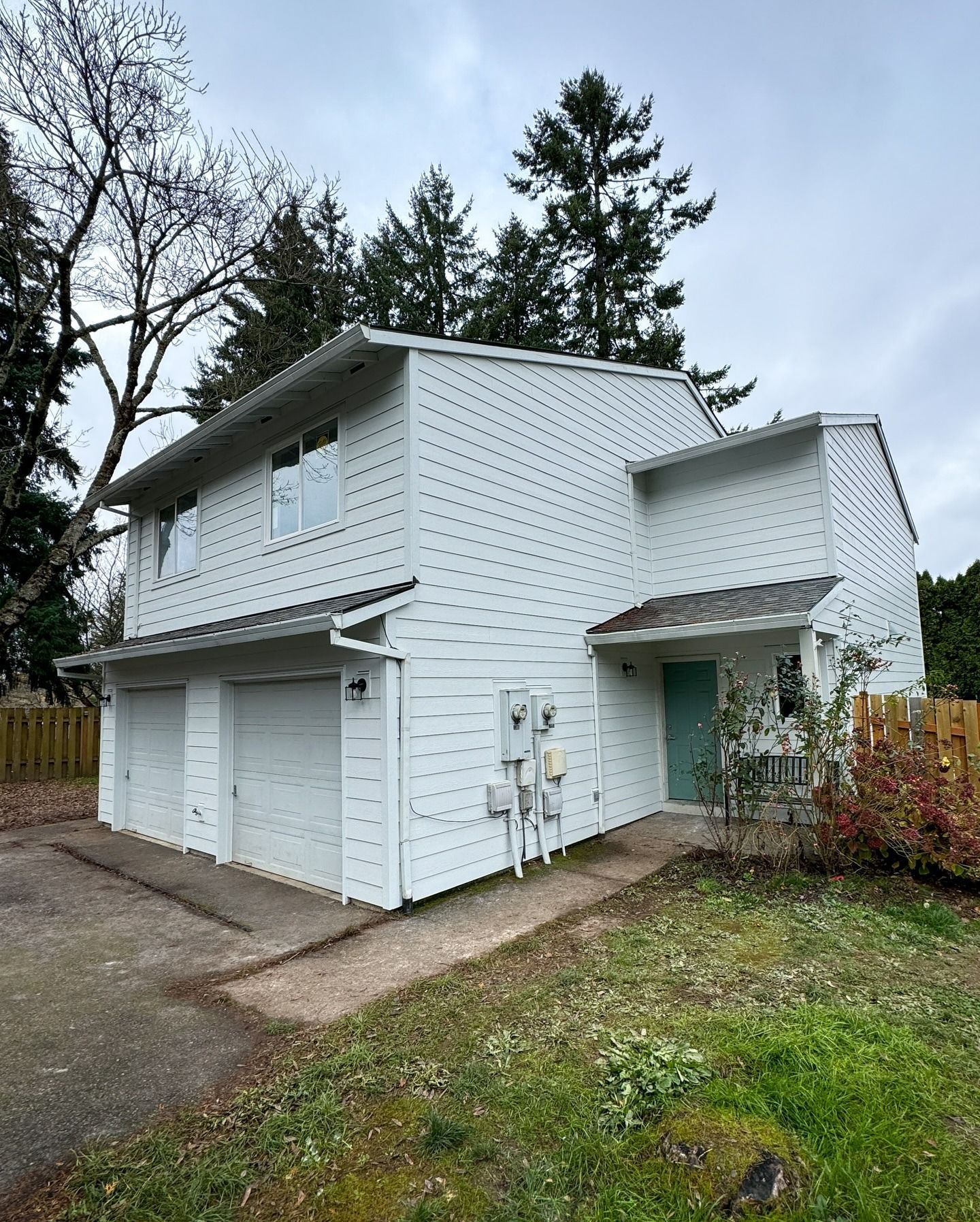 A two-story, white-sided duplex with two garage doors and a light green front door, surrounded by trees and a lawn.