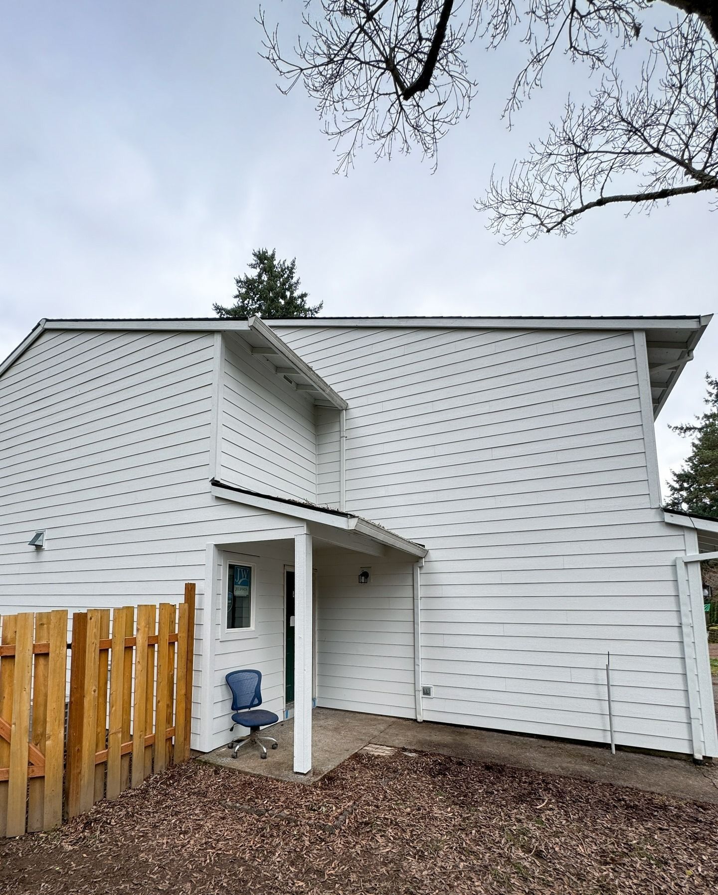 A two-story white house with light-colored siding, a small covered entryway, and a wooden fence on the left side.