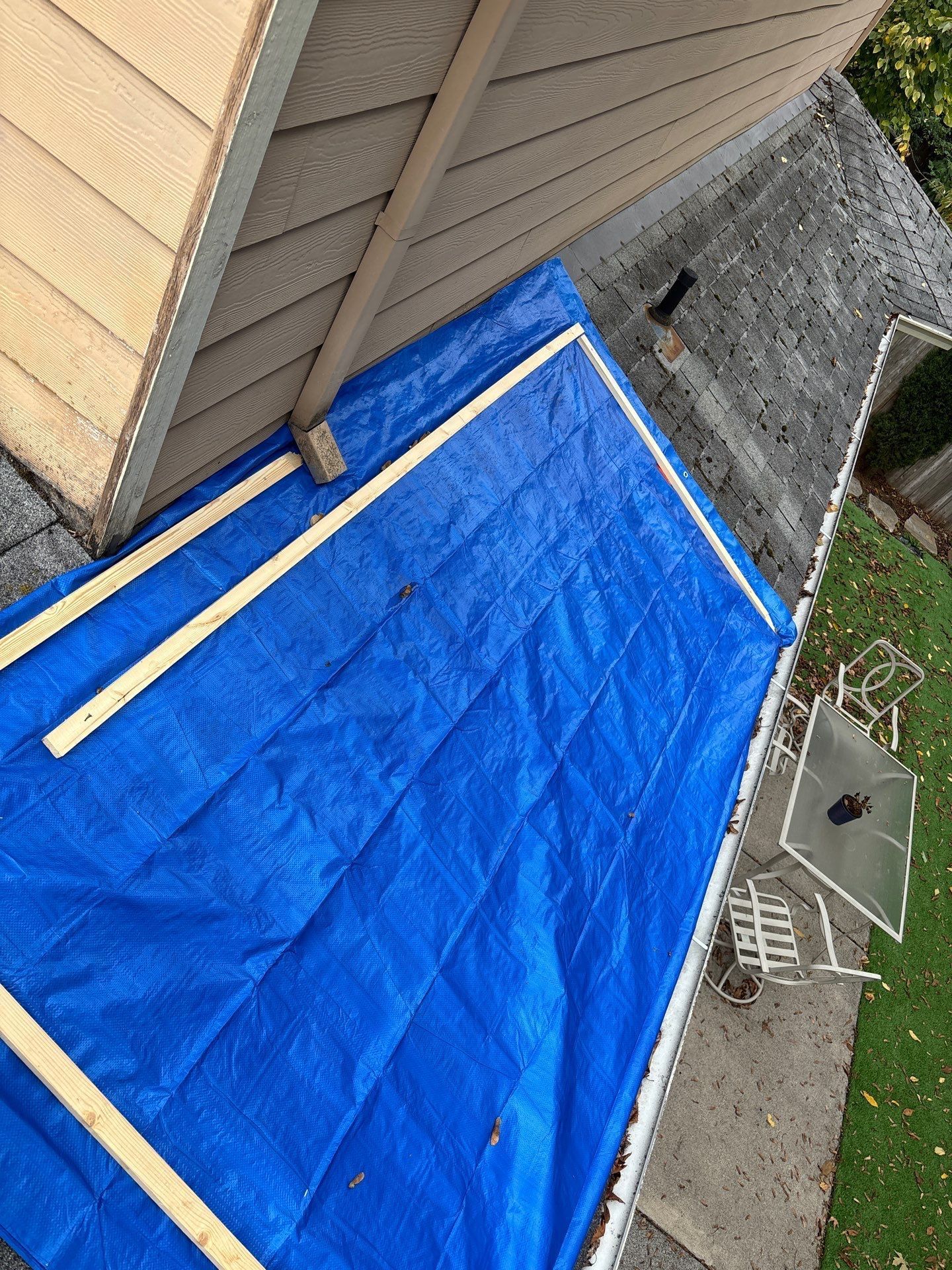 A blue tarp covers a section of a shingled roof, secured with wooden boards along the edges near a house wall.