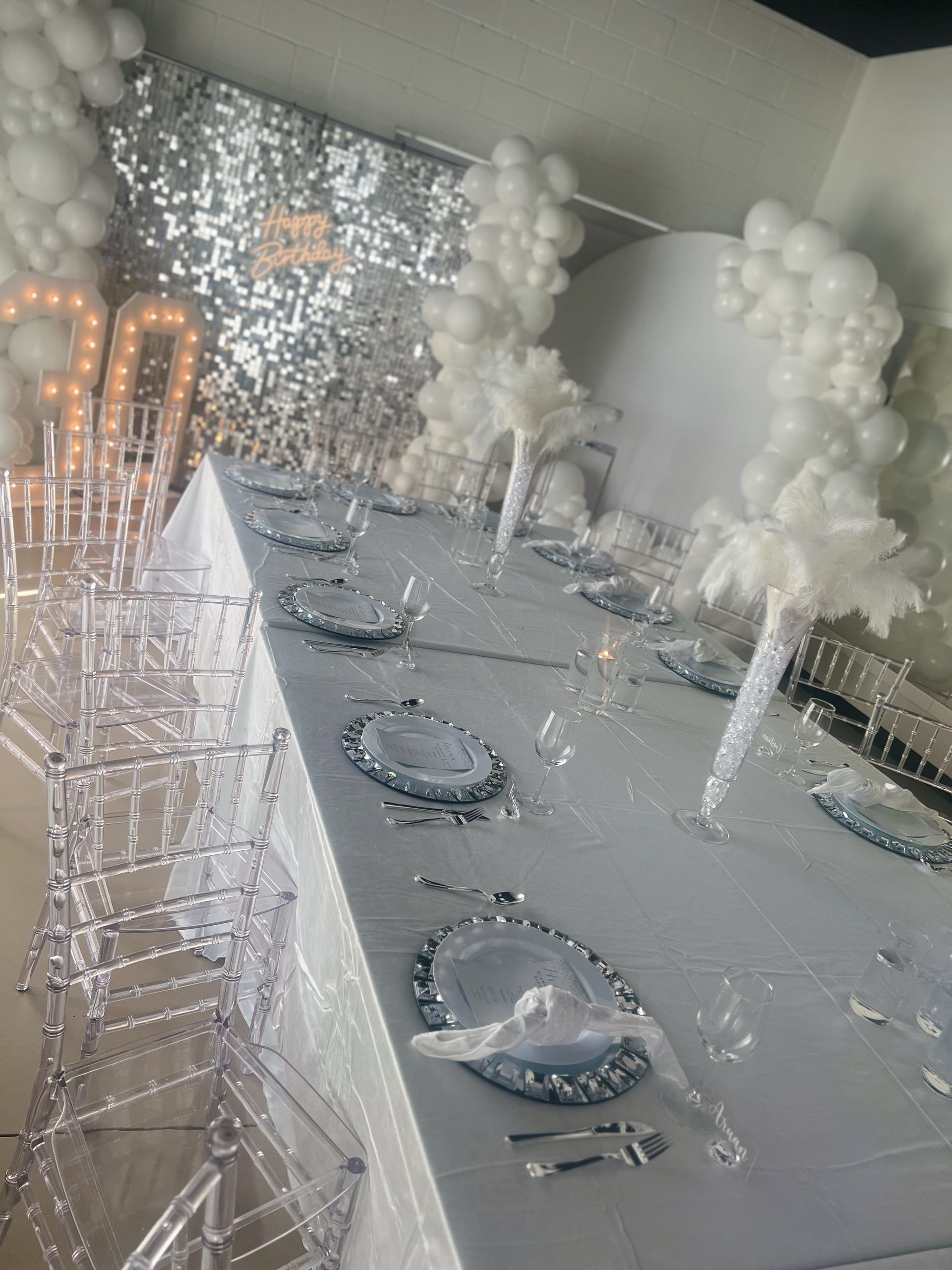 A long table with plates , glasses , silverware and balloons on it.