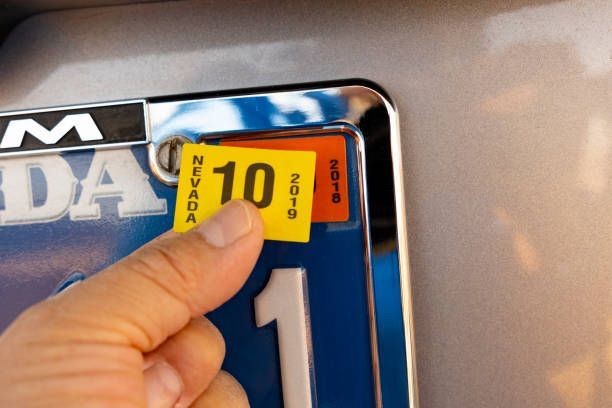 Hand placing a 2019 Nevada license plate sticker (yellow) on a car's license plate.