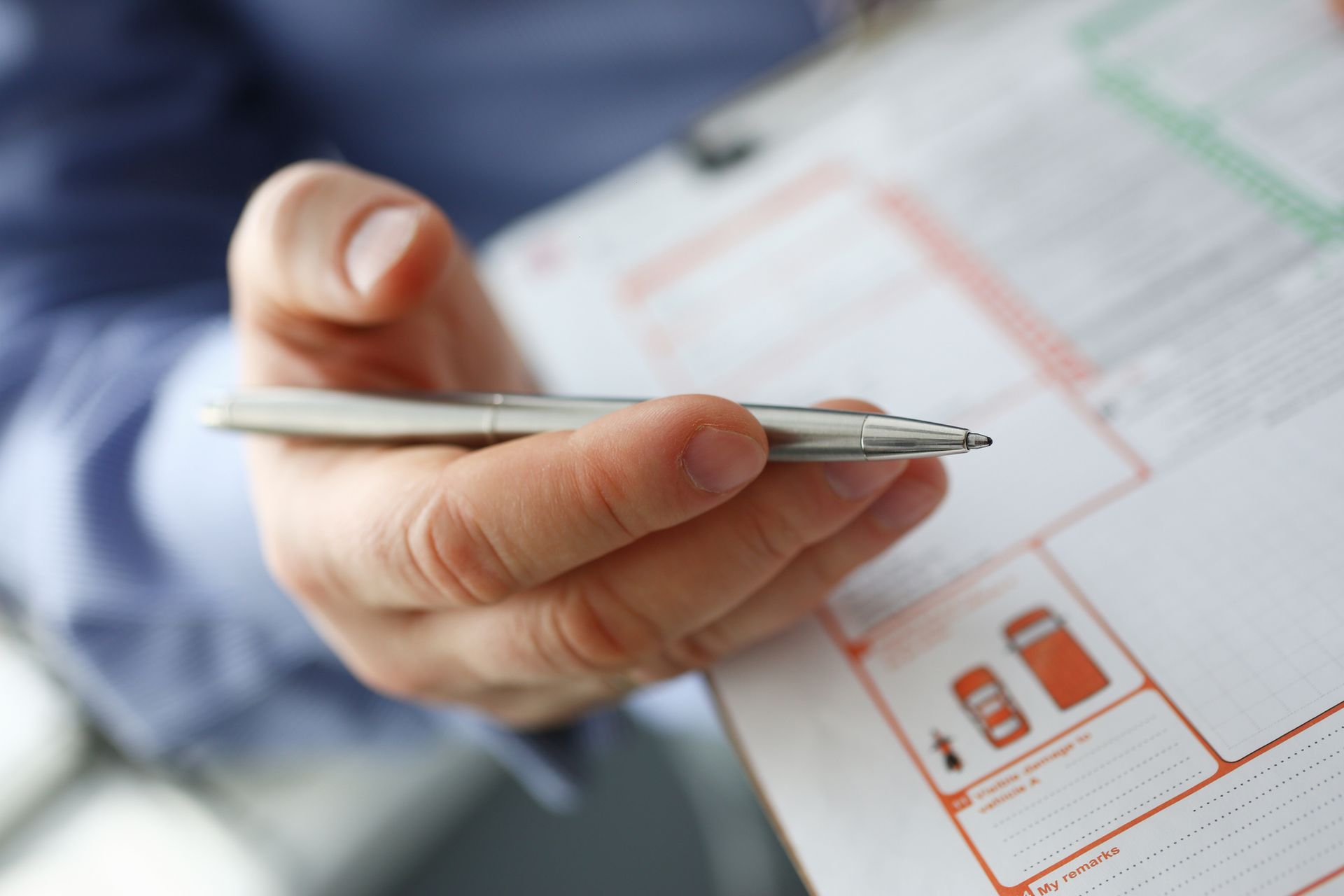 Person holding a silver pen over a clipboard with orange and green markings; blurred background.